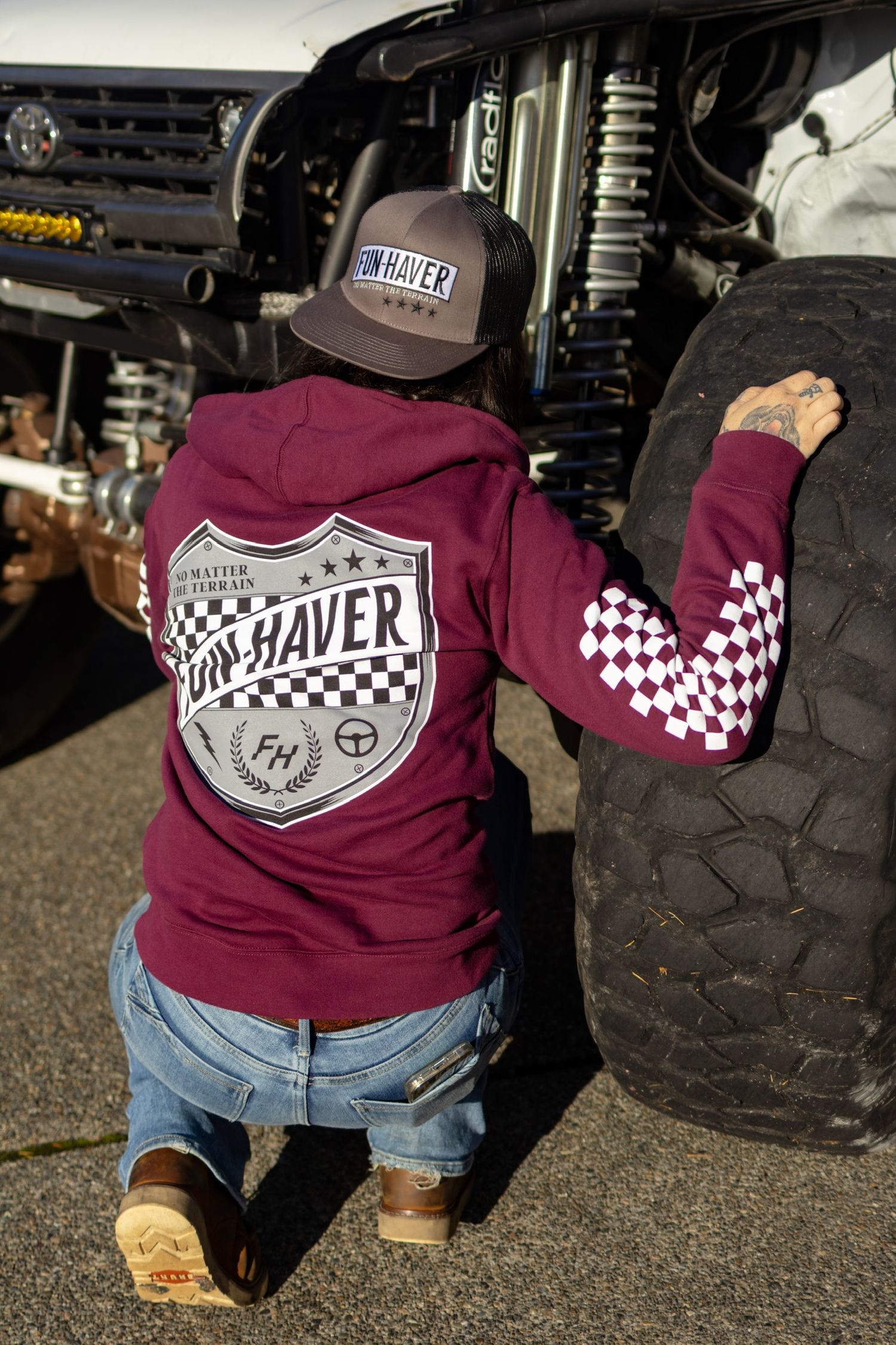 Burgundy sweatshirt with racing shield logo and checkered pattern displayed near off-road tires.