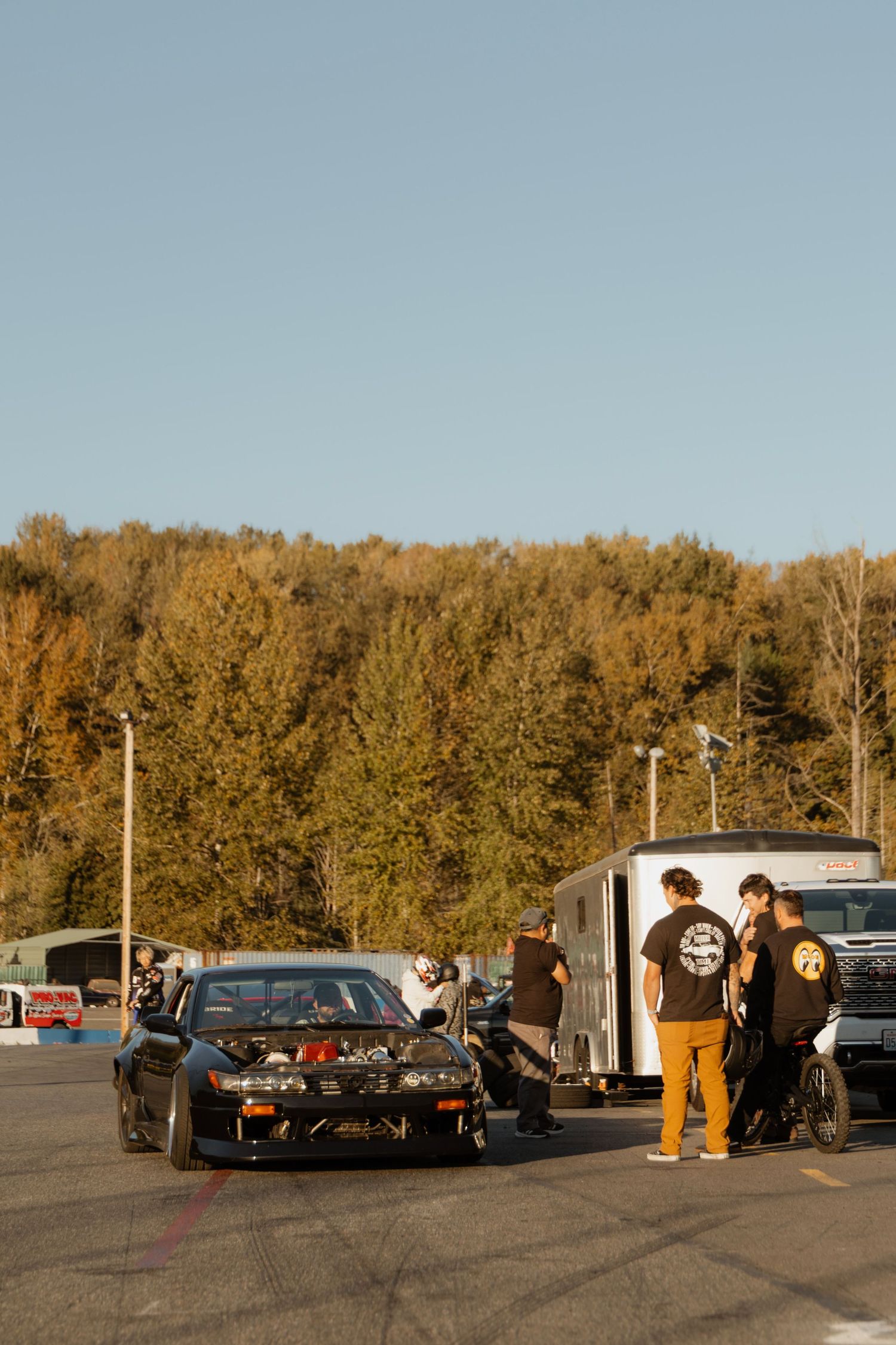 People gathered in a parking lot with cars and autumn trees in the background during golden hour.