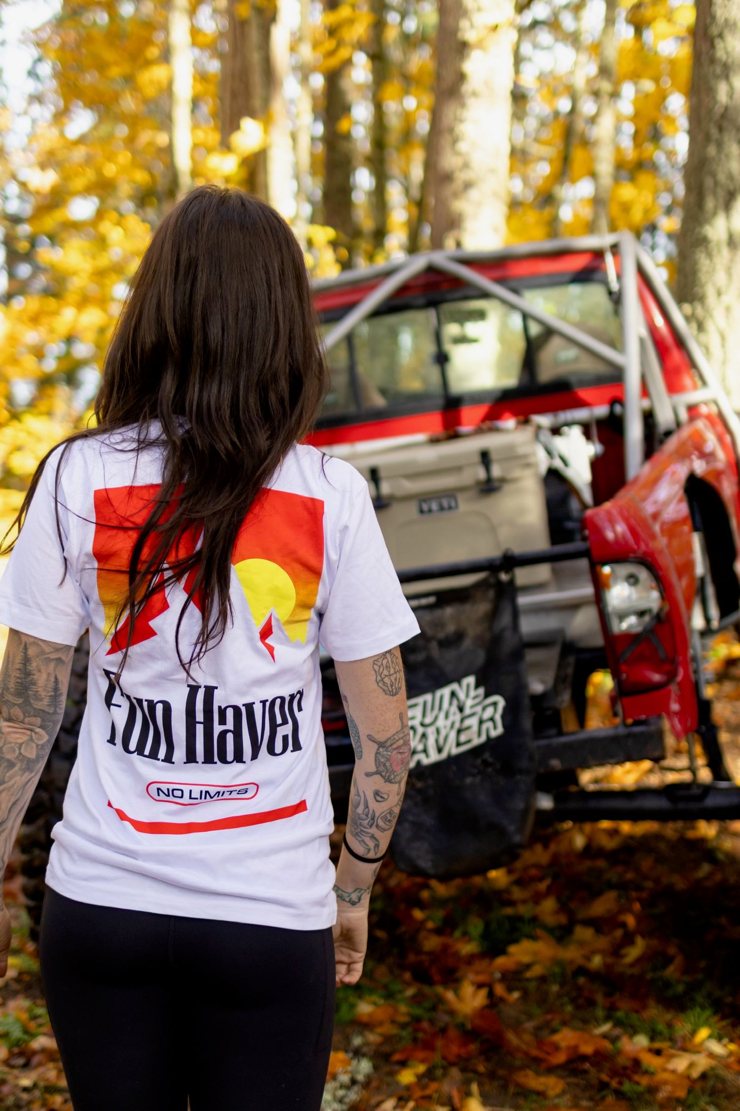 White t-shirts with outdoor mountain designs displayed in autumn forest setting with off-road vehicles in background.
