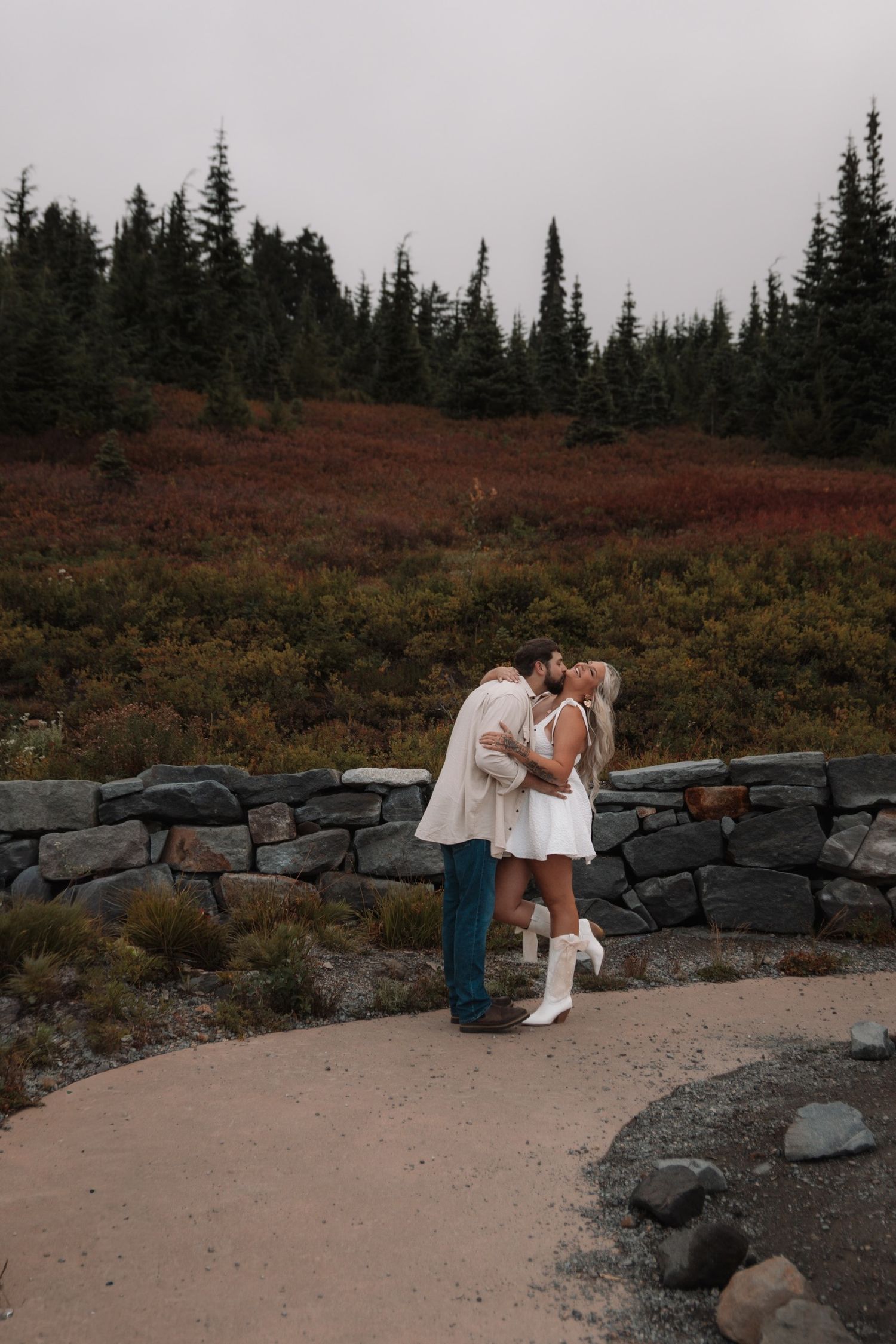 Couple poses romantically on a mountain path with stone wall and evergreen forest backdrop.