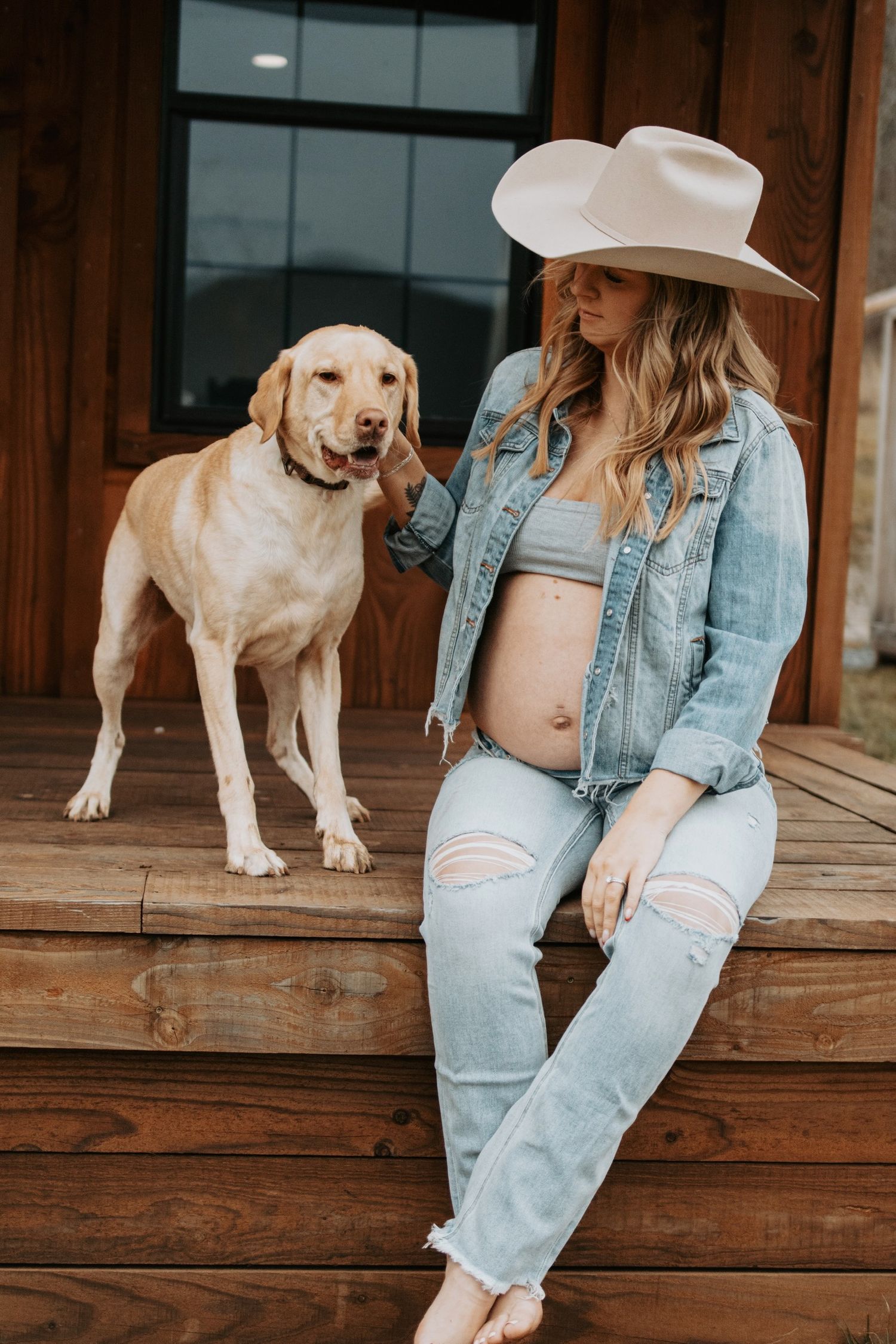Pregnant woman in western wear sits with yellow Labrador on wooden porch.