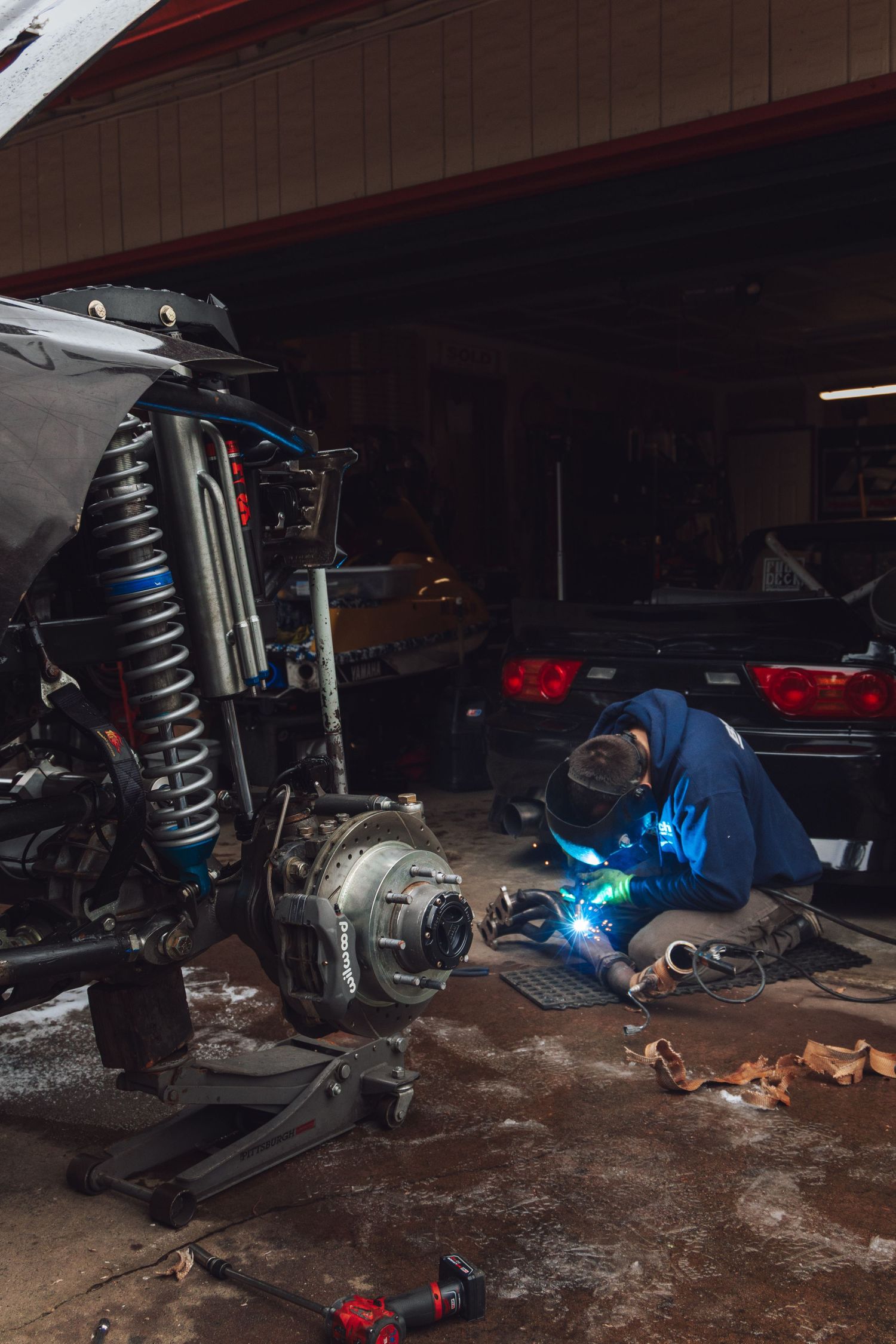 Vehicle suspension parts and welding work being done in dark garage setting.