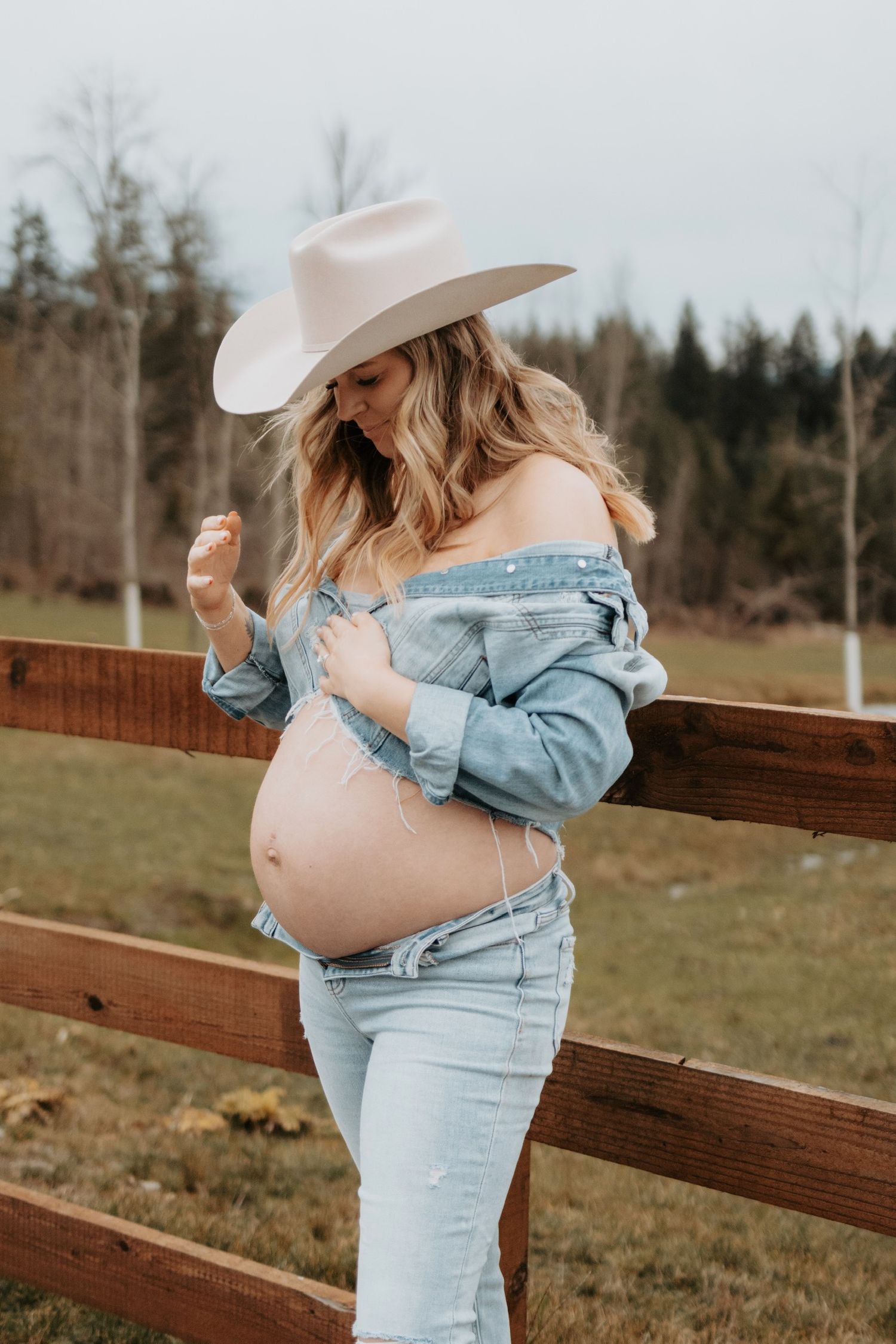 Rustic maternity photo with woman in denim and white cowboy hat leaning on fence.