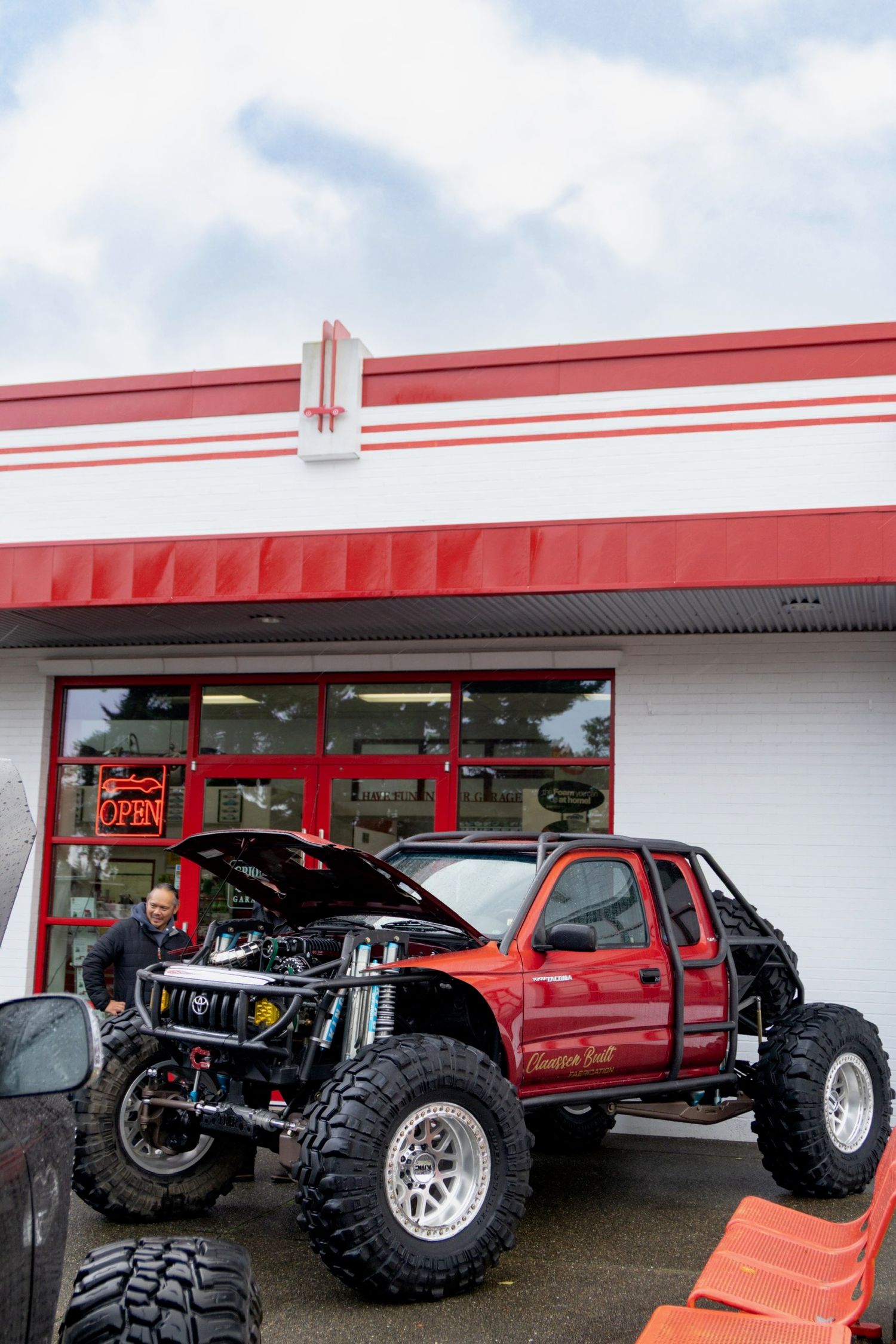 Line of lifted trucks with large off-road tires parked in front of a commercial building with red trim.