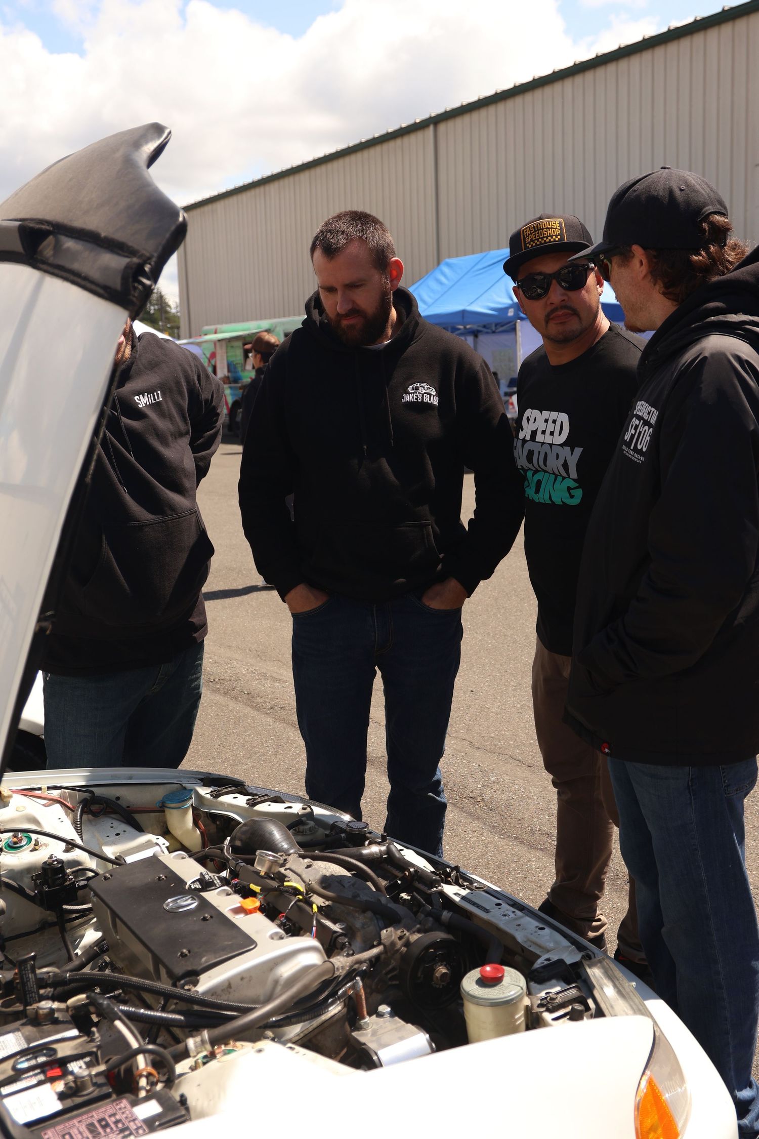 A group of people gathered around examining an open vehicle engine compartment in a workshop setting.