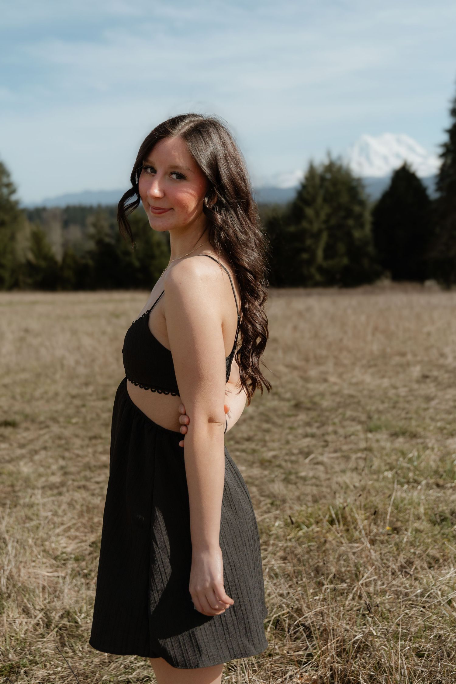 A person in a black dress stands in a golden field on a sunny day with mountains visible in the distance.