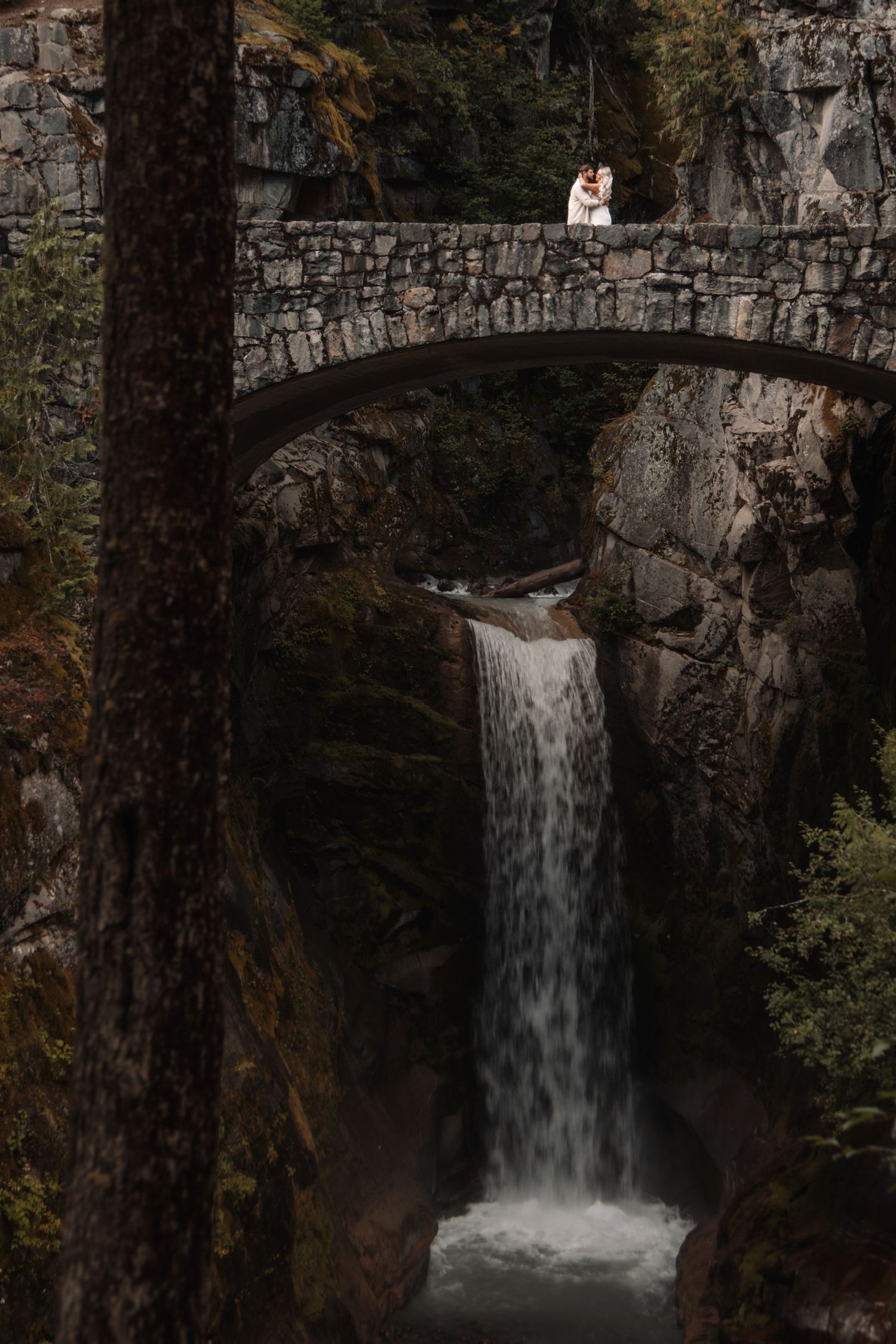 A scenic waterfall cascades beneath a stone bridge in a forest setting during autumn.