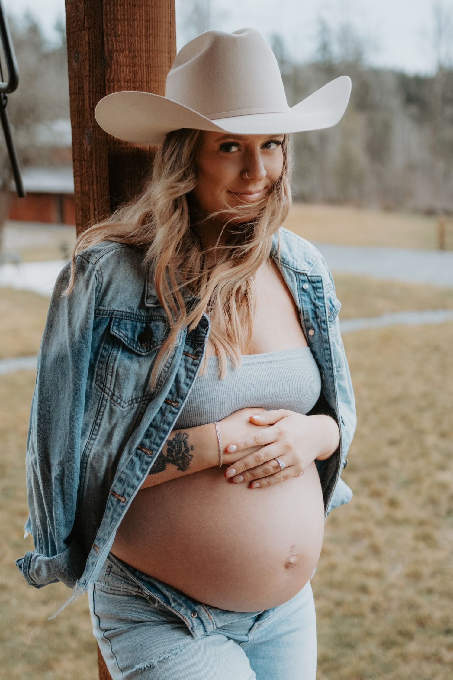Maternity portrait with cowboy hat and denim jacket against rustic backdrop.