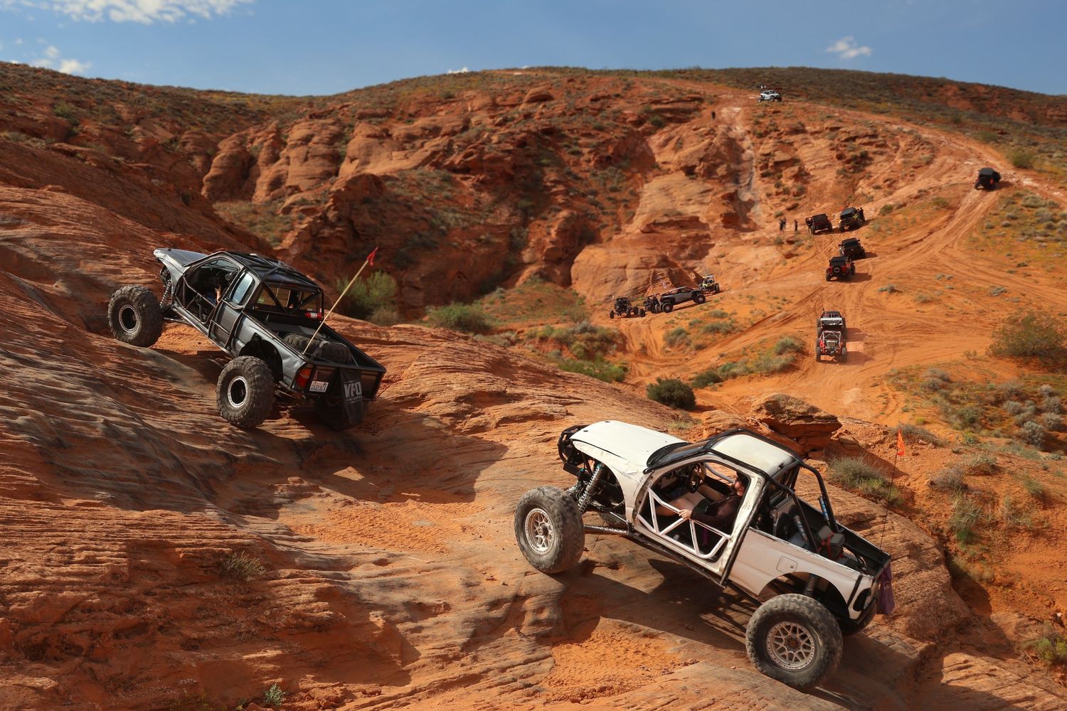 Off-road vehicles navigate challenging red rock terrain on a desert trail under blue skies.