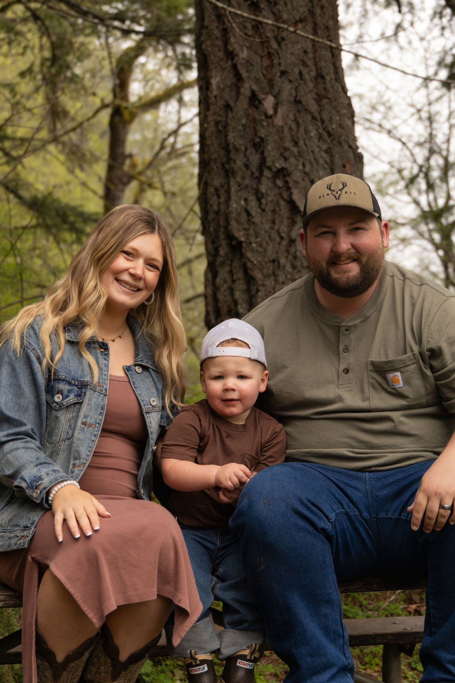 A family sits together on a log in a wooded area wearing casual clothing and smiling at the camera.