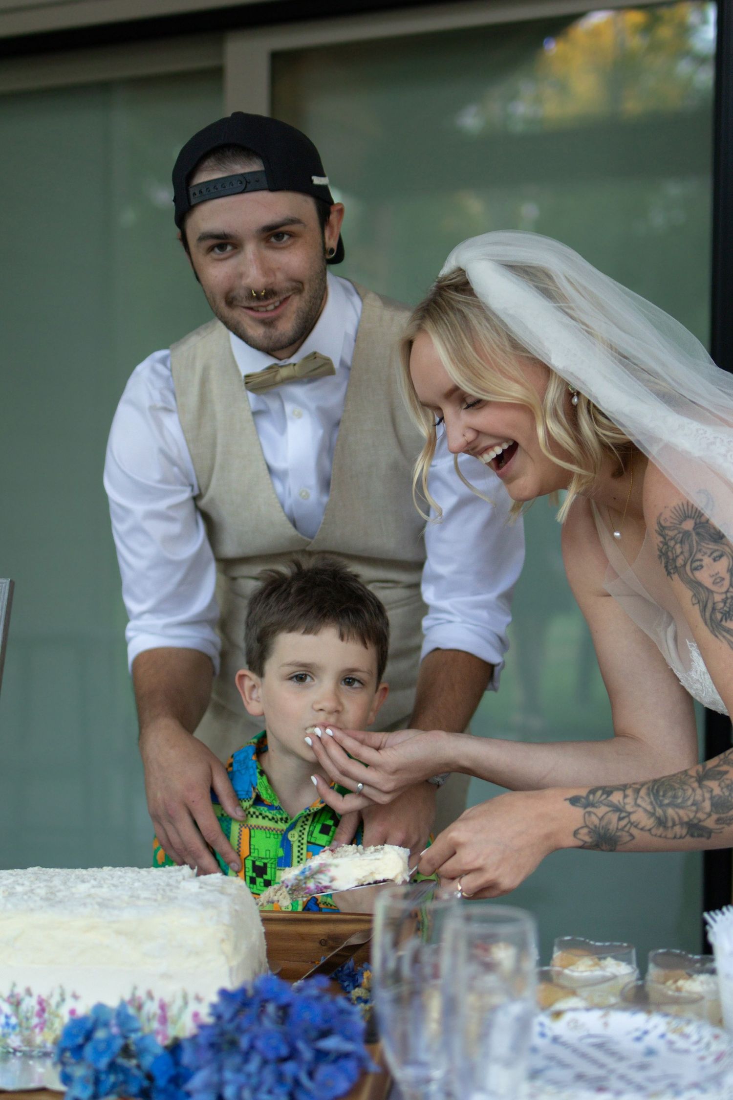 A joyful wedding moment at the cake table as a couple shares dessert with a young child in a casual outdoor setting.
