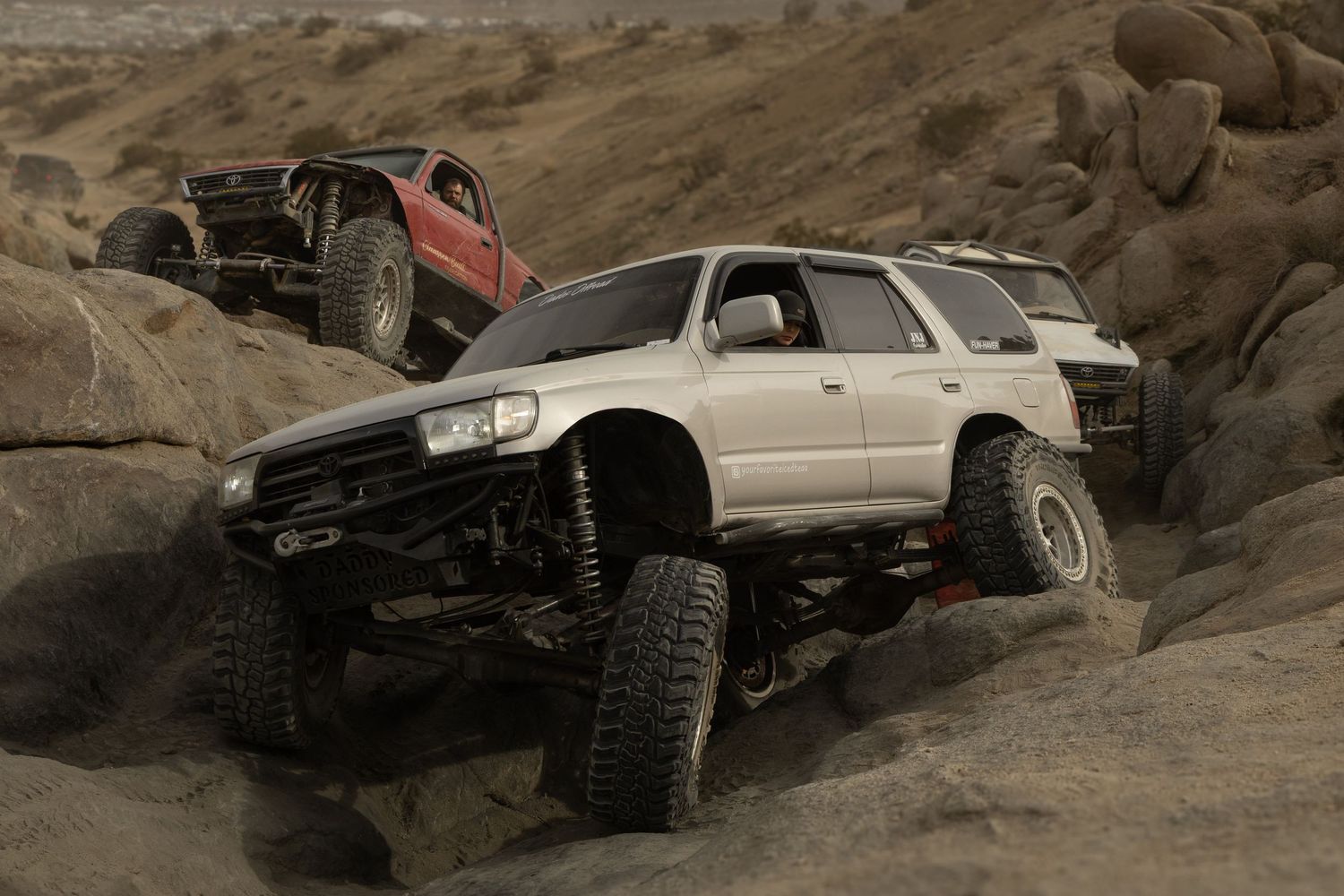 Modified Toyota 4Runner with large off-road tires climbing over rocky terrain with another vehicle visible in background.