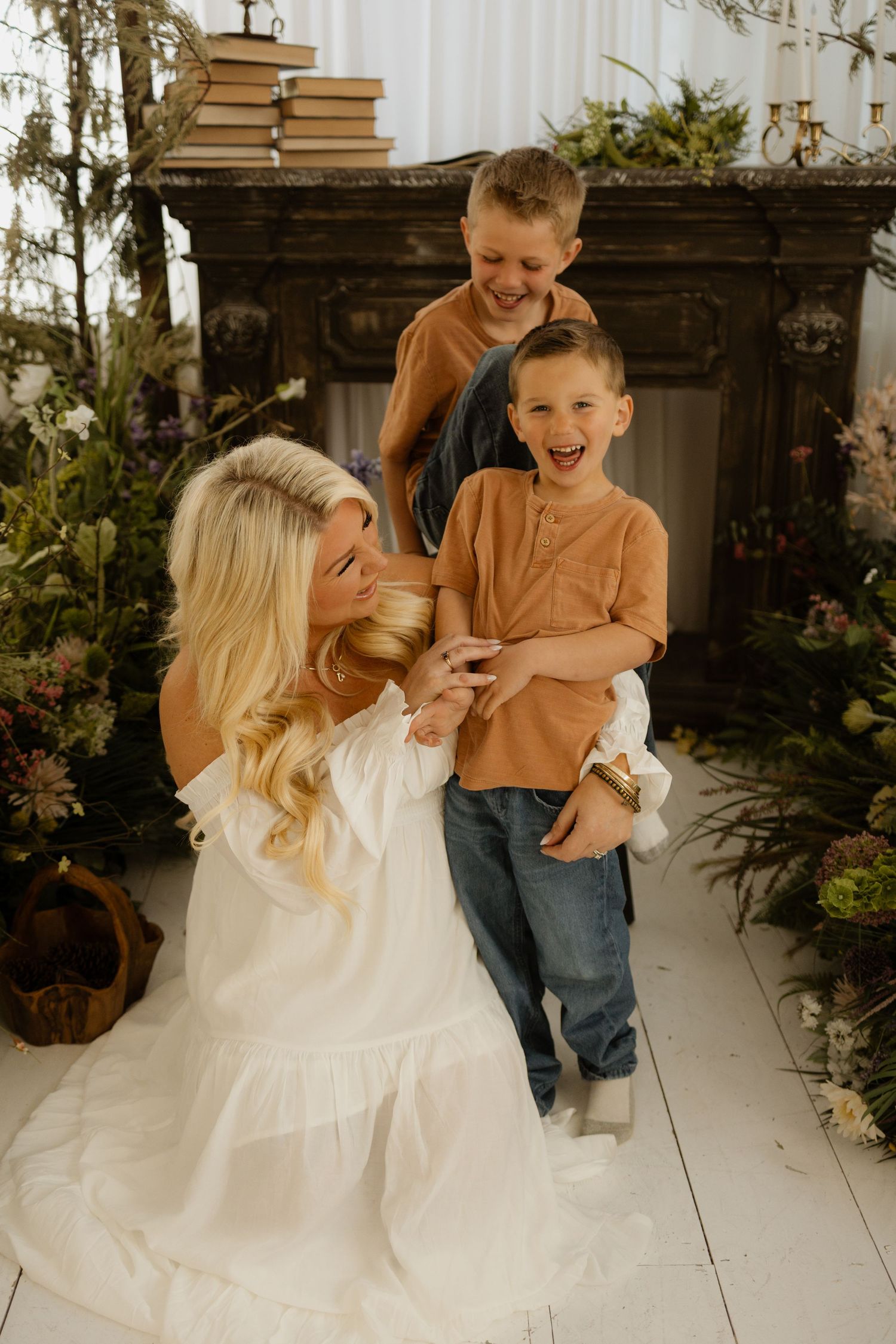 A blonde woman in a white dress laughs with two boys in matching brown shirts surrounded by lush florals.