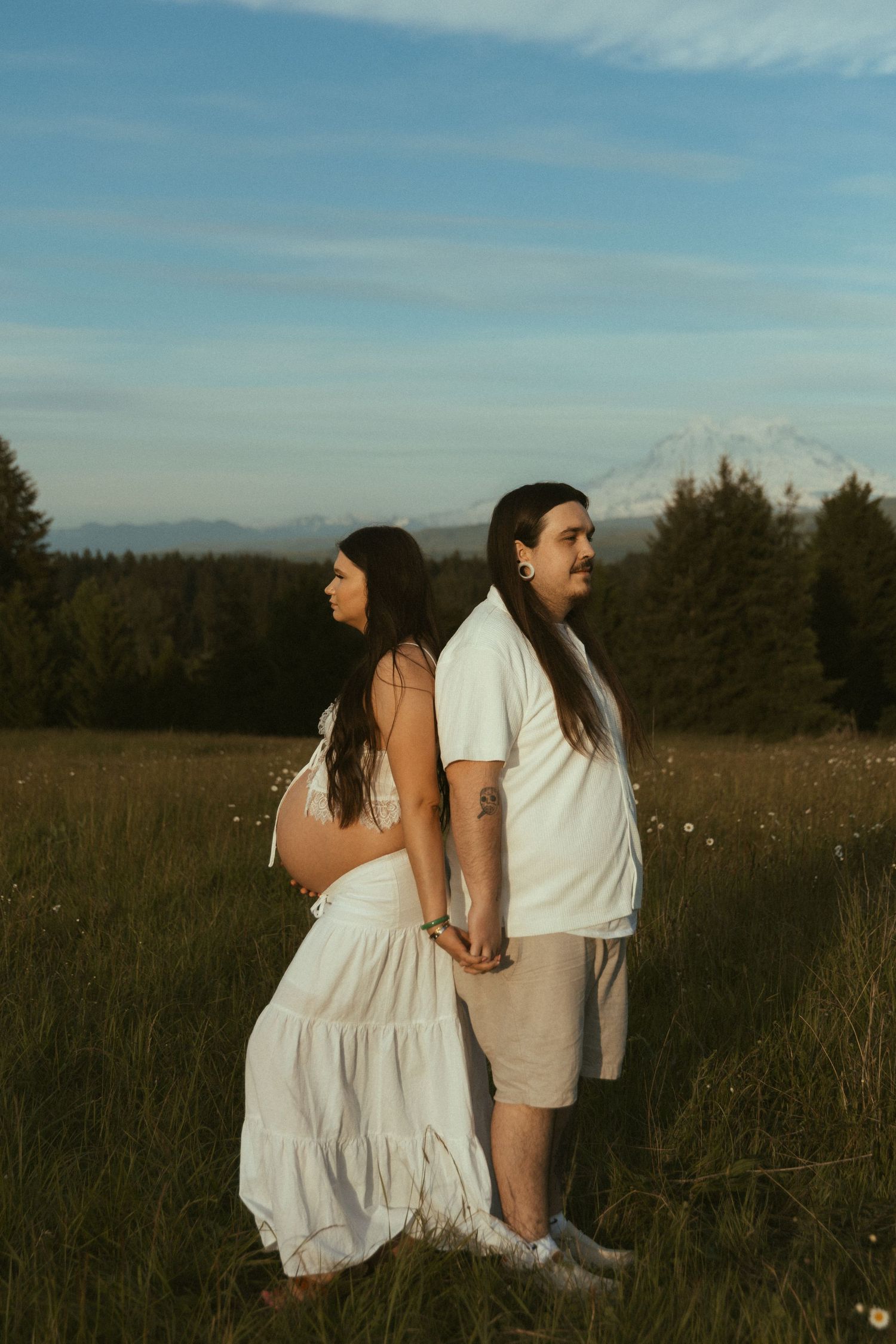 Pregnant couple holding hands in a sunlit meadow, woman in white dress, man in white shirt, with mountain backdrop.