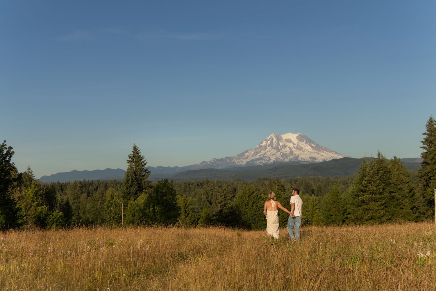 Two figures in white stand in a golden meadow with Mount Hood rising majestically in the distance.
