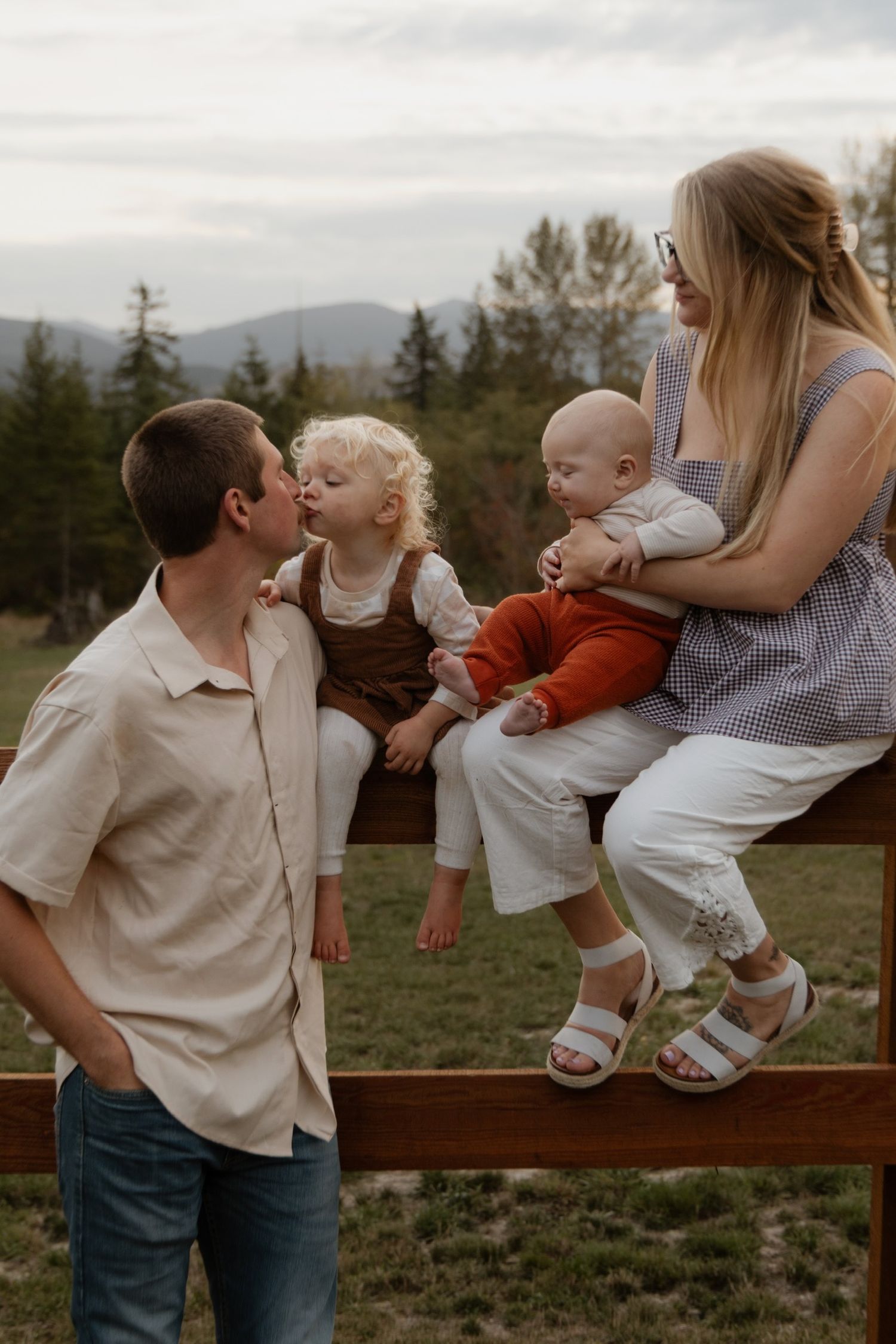 A family shares playful moments while sitting on a wooden fence in a rural setting.