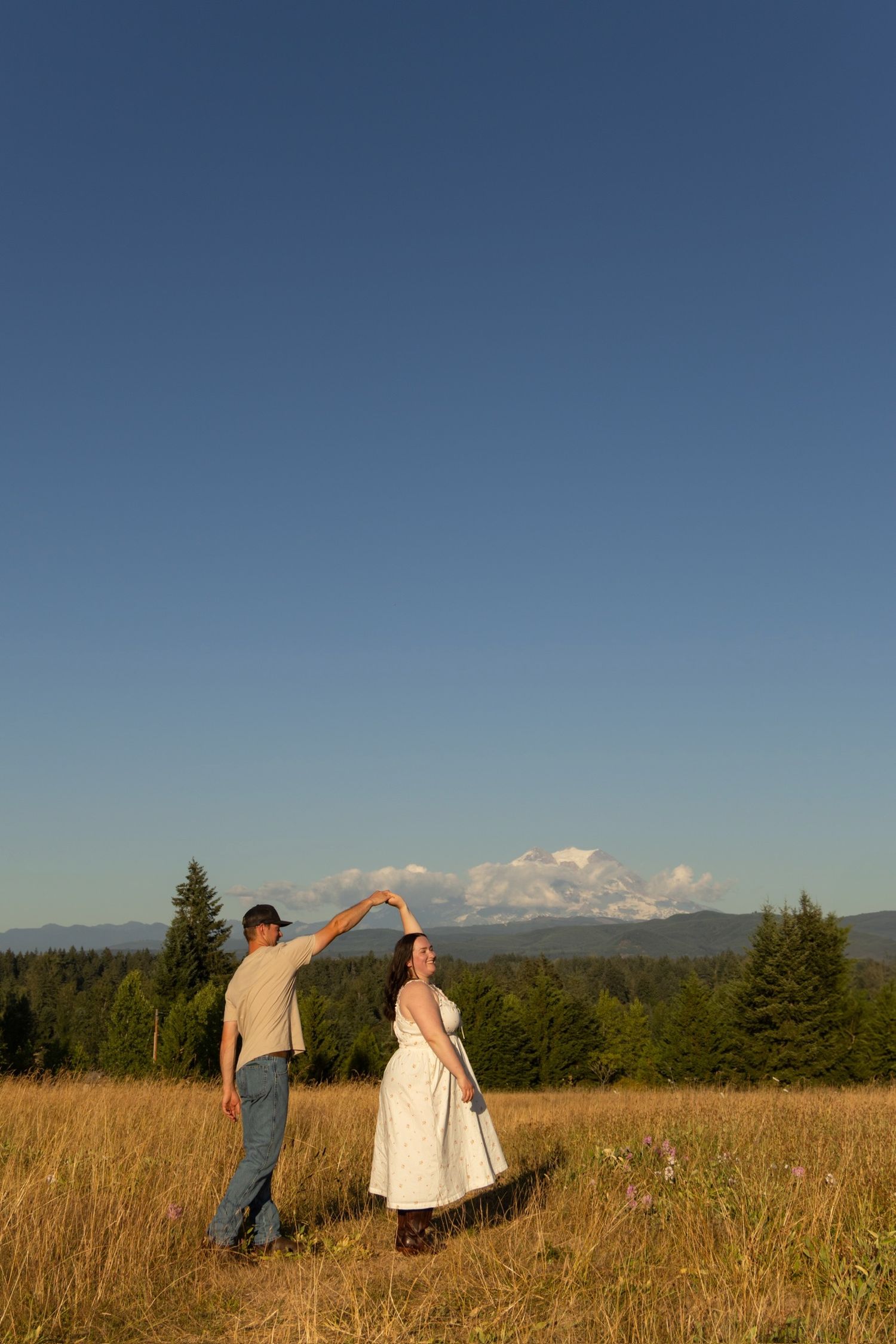 Two people stand in a golden field with mountains and evergreen trees visible in the distance against a blue sky.