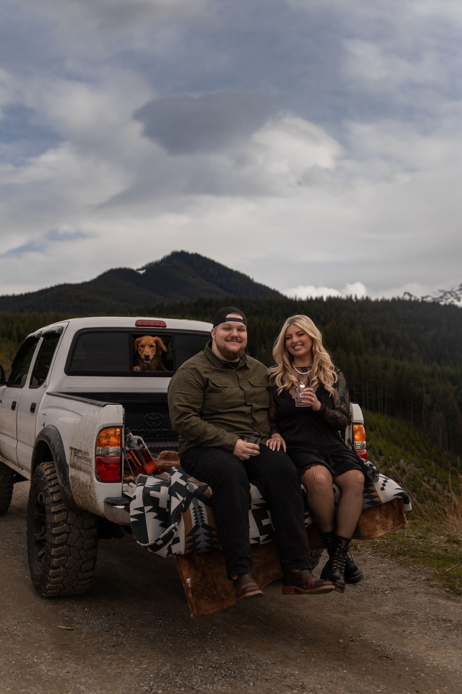 Couple sitting with their dog by a pickup truck in front of mountain scenery.