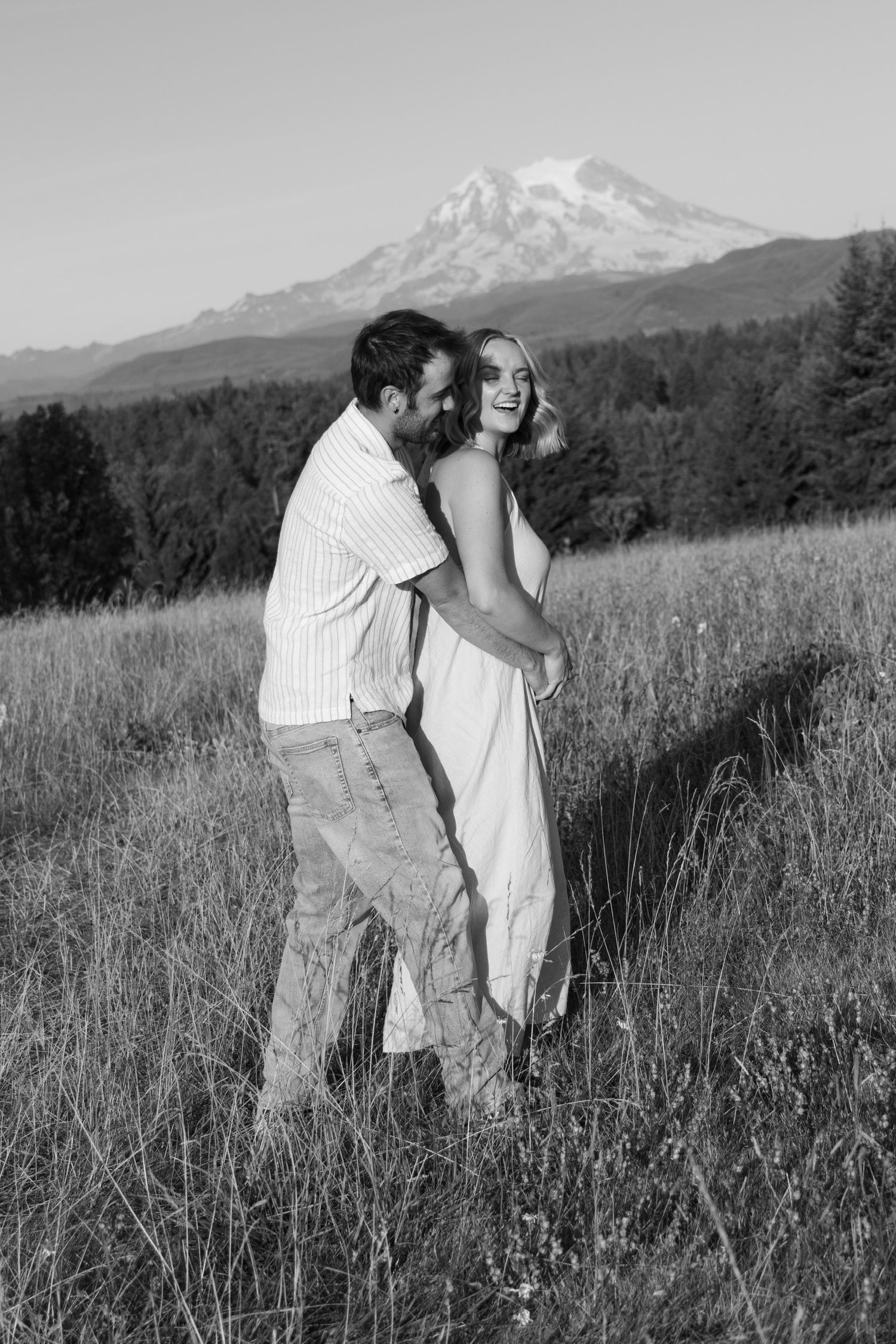 Black and white photo of a couple embracing in a field with mountains in the background.