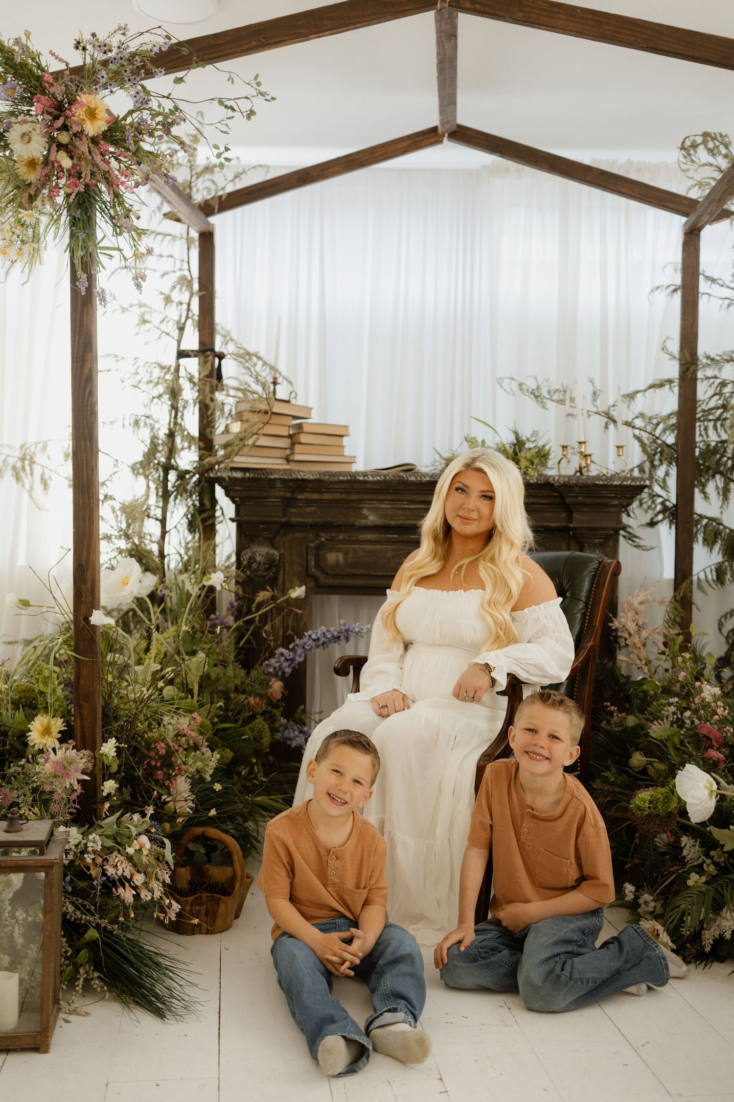 A woman in a white dress poses with two young boys amid lush floral arrangements under a wooden arch.