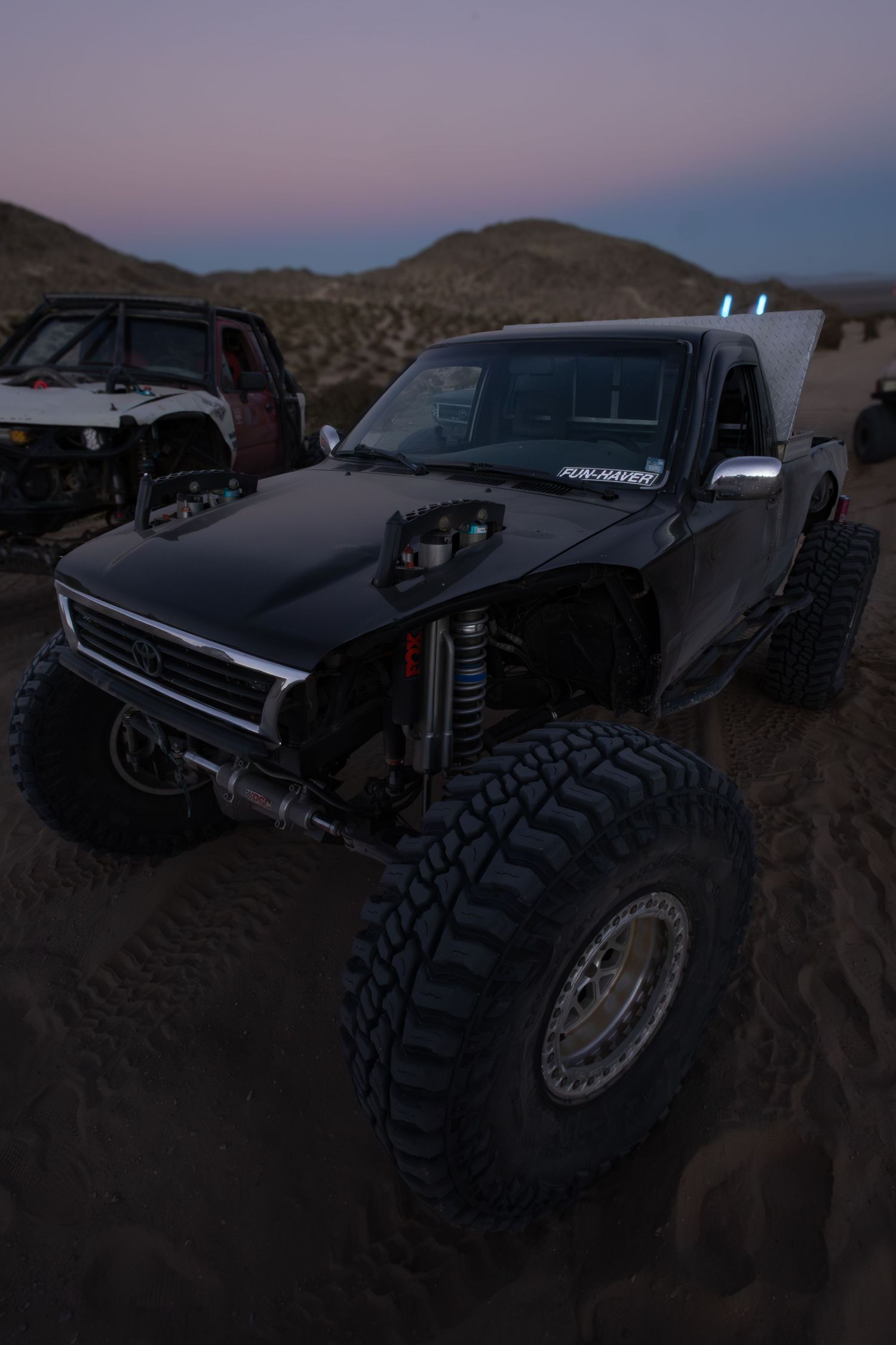 Modified black off-road truck with large rugged tires and lifted suspension sits in desert terrain at dusk.