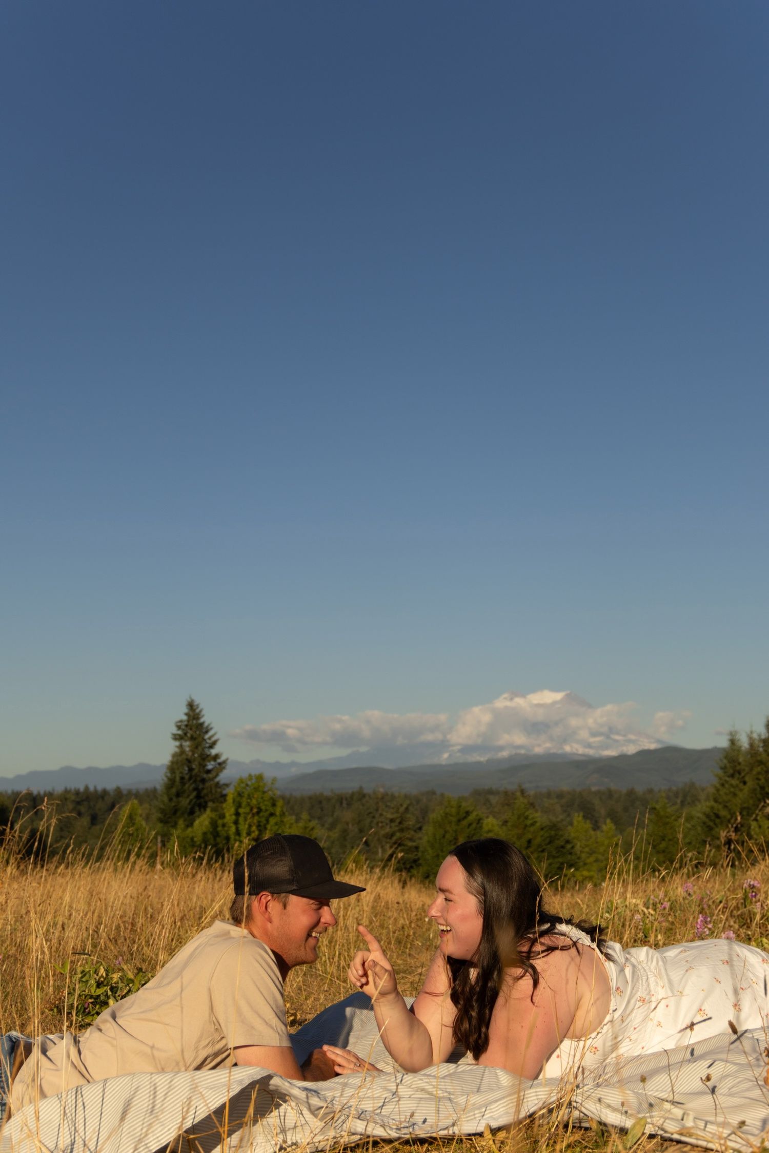 Couple lying in tall grass field with mountains and blue sky in background.