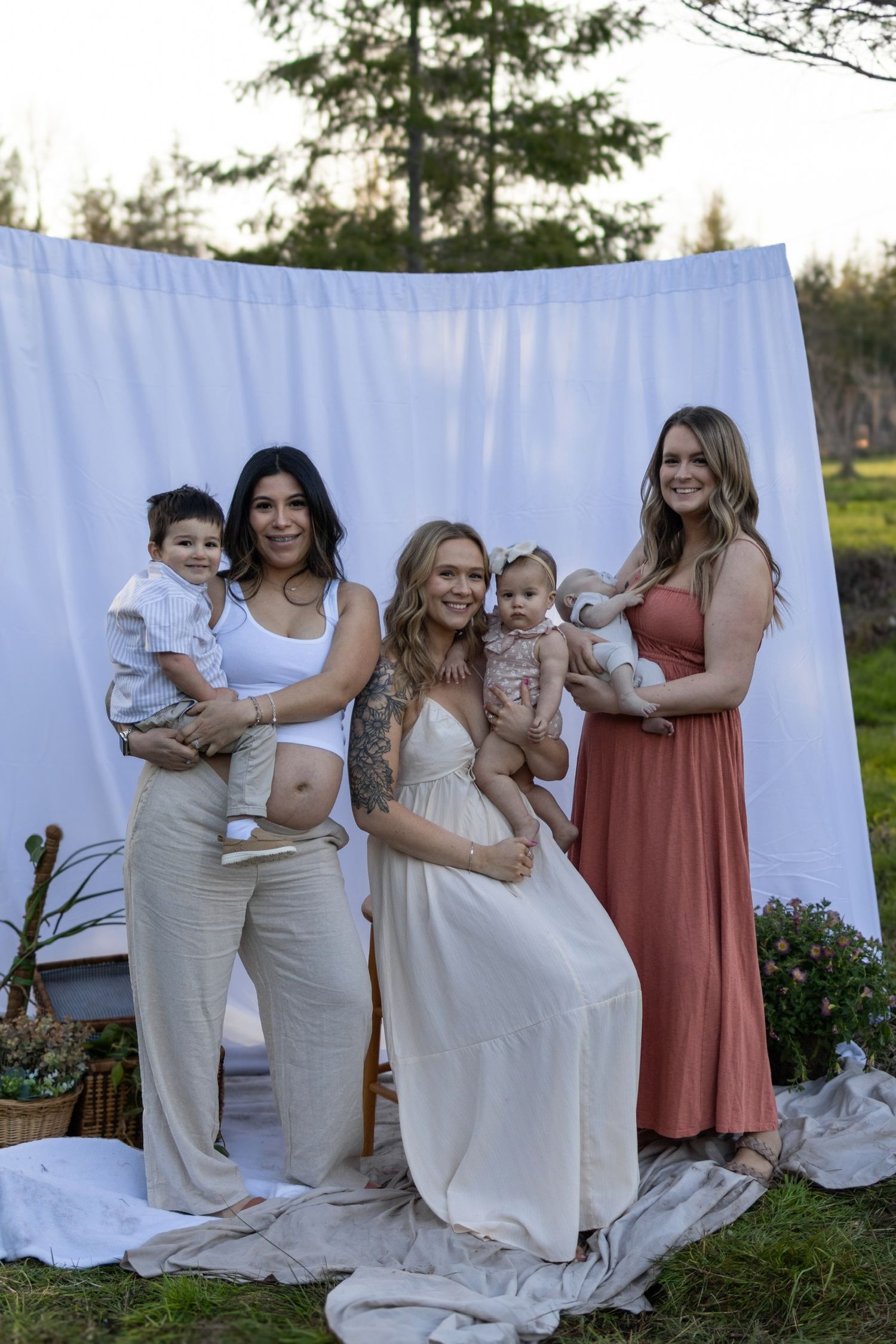 A group of people in white and pink dresses pose together against a white sheet backdrop outdoors.