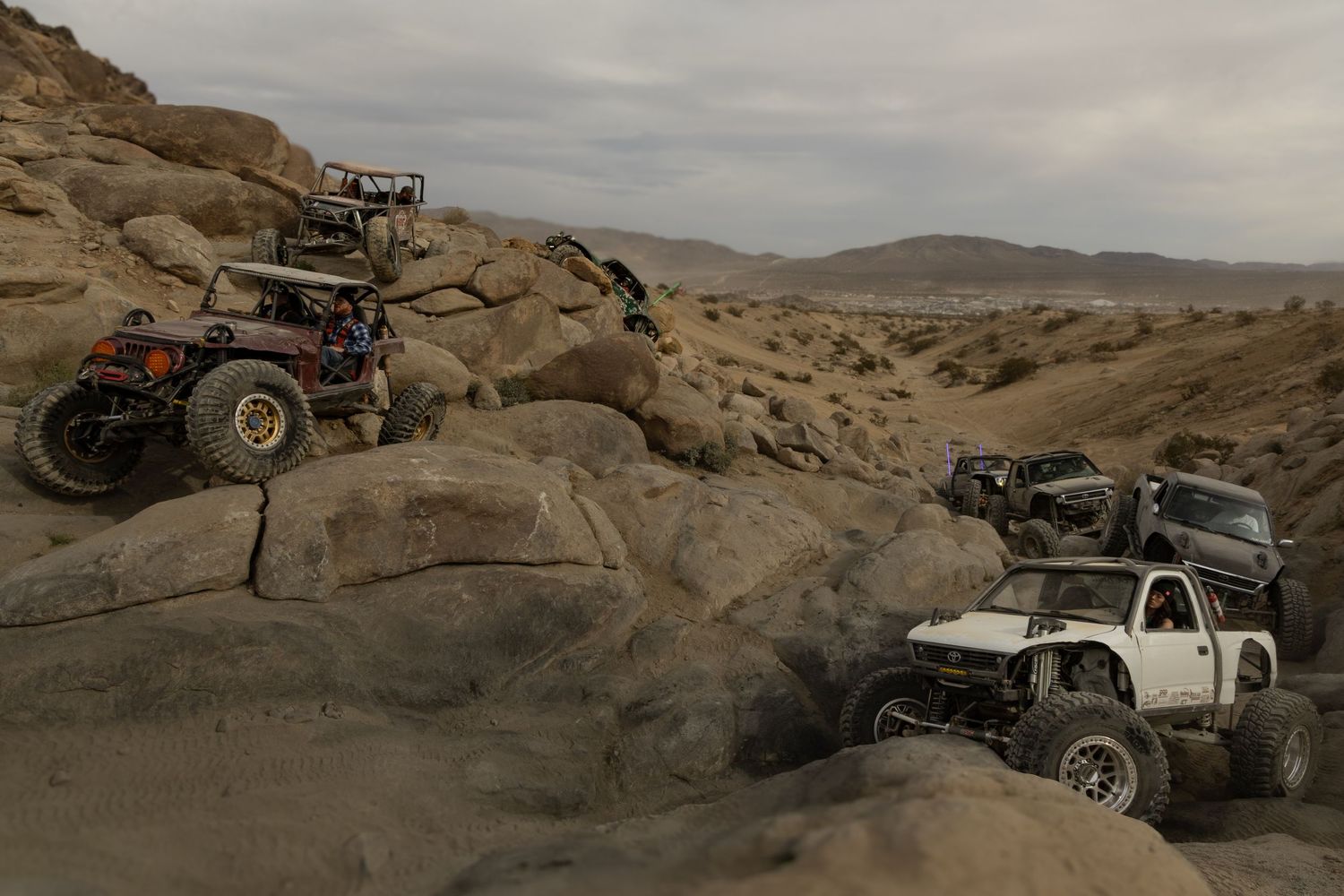 Off-road vehicles traversing rocky desert terrain under cloudy skies.