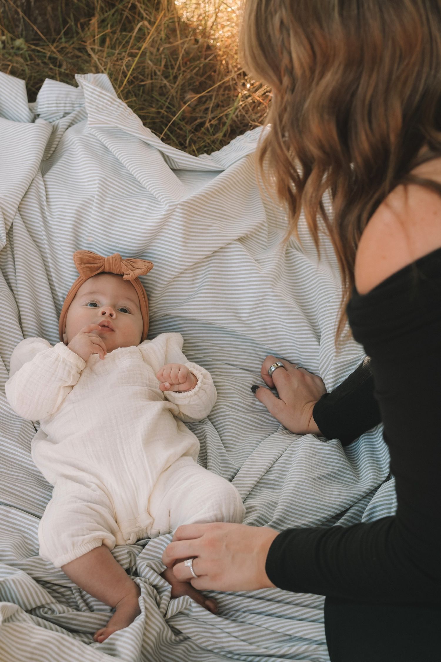 Baby in white outfit lying on striped blanket during outdoor photo session.