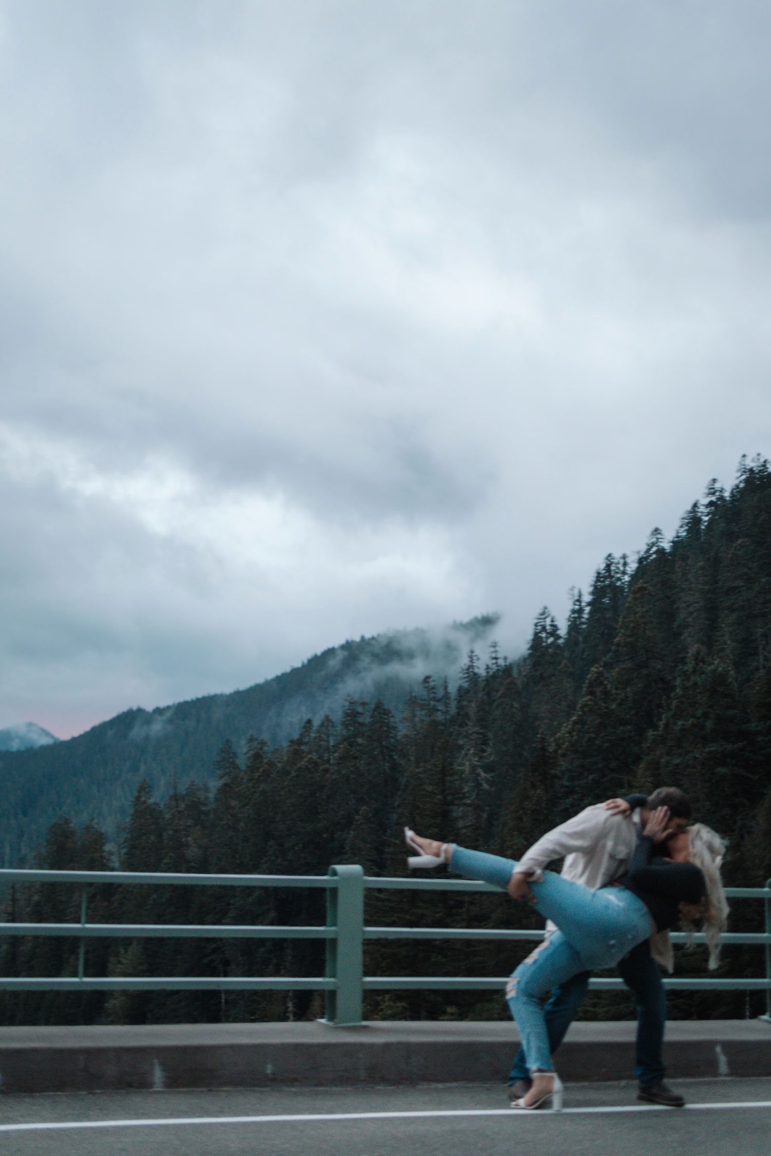Two people share a romantic moment on a bridge railing with misty mountain forest scenery in the background.