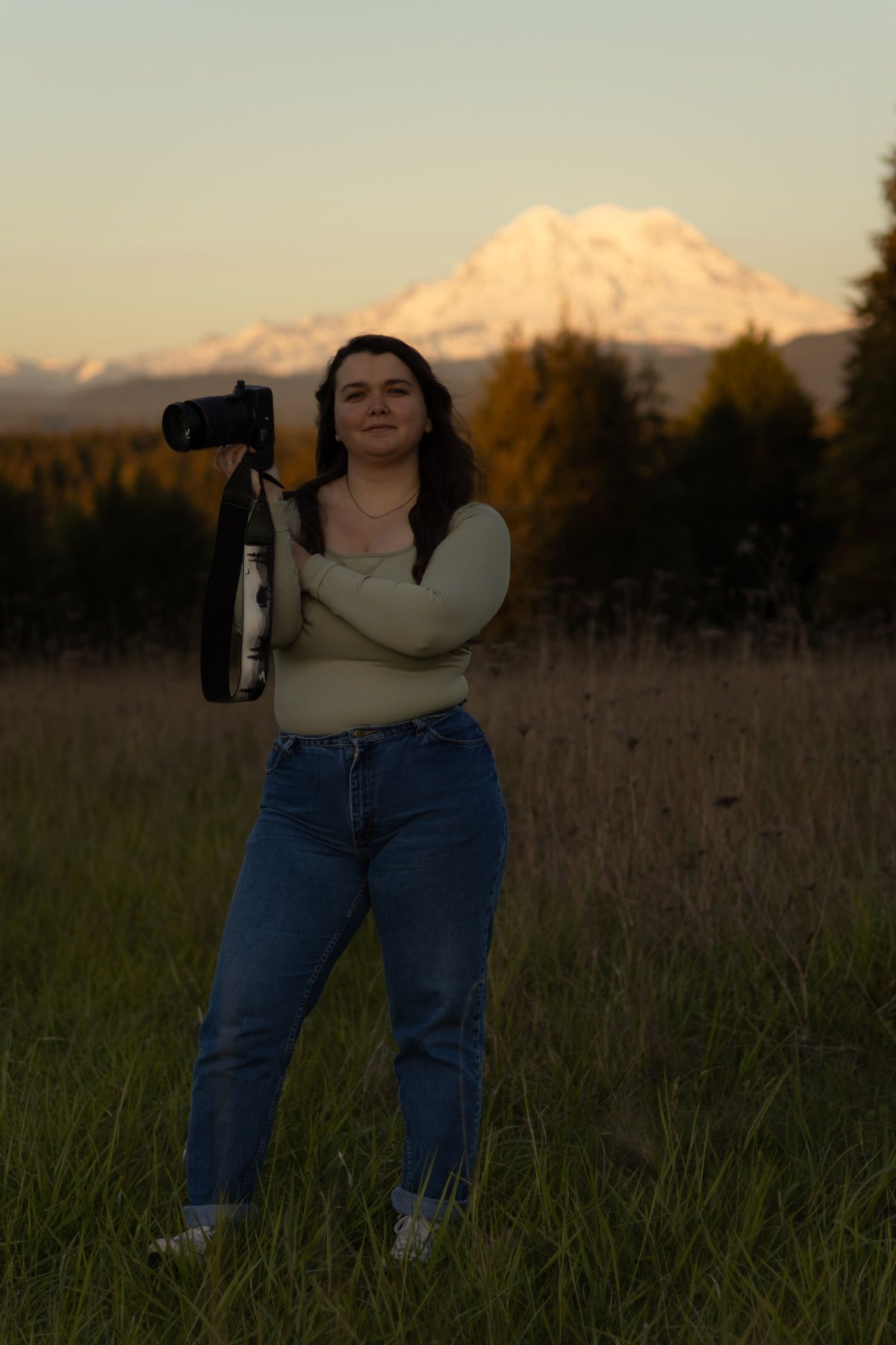 Individual in light sweater and jeans stands in field with snow-capped mountains visible at sunset.