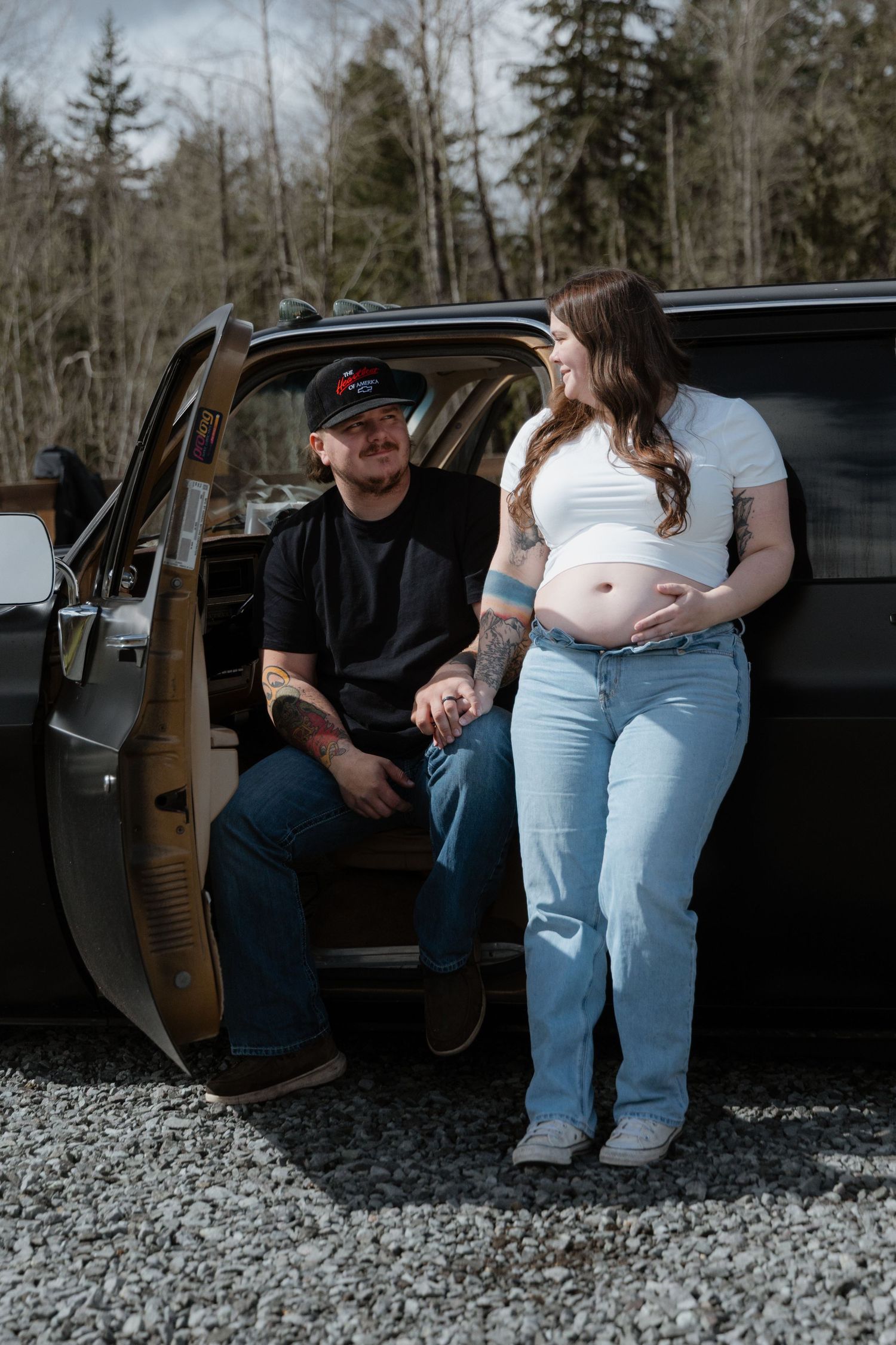 A couple poses by a black truck on gravel, with the pregnant woman showing her belly while standing beside the open door.