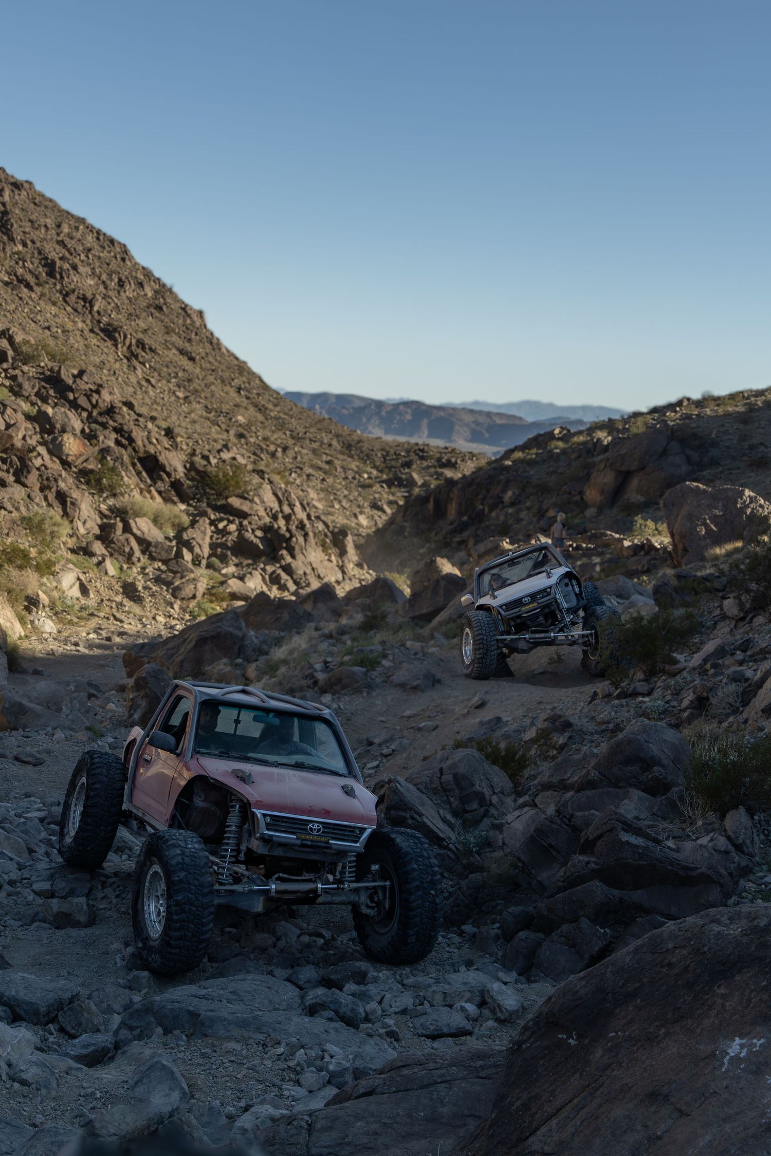 Two off-road vehicles navigate a rocky desert trail through steep canyon terrain under clear blue skies.