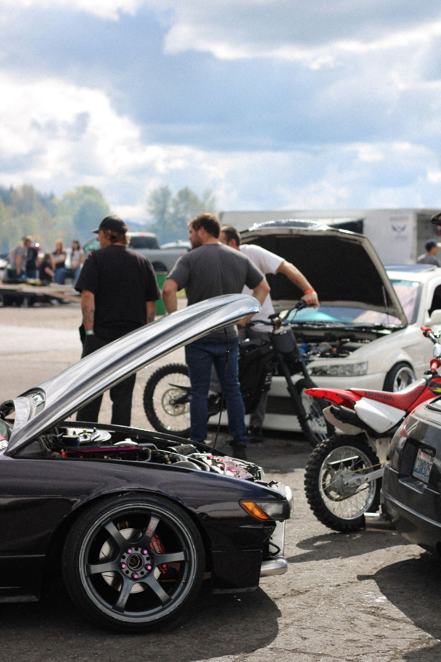A row of luxury sports cars and motorcycles on display at an outdoor car show with their hoods open for viewing.