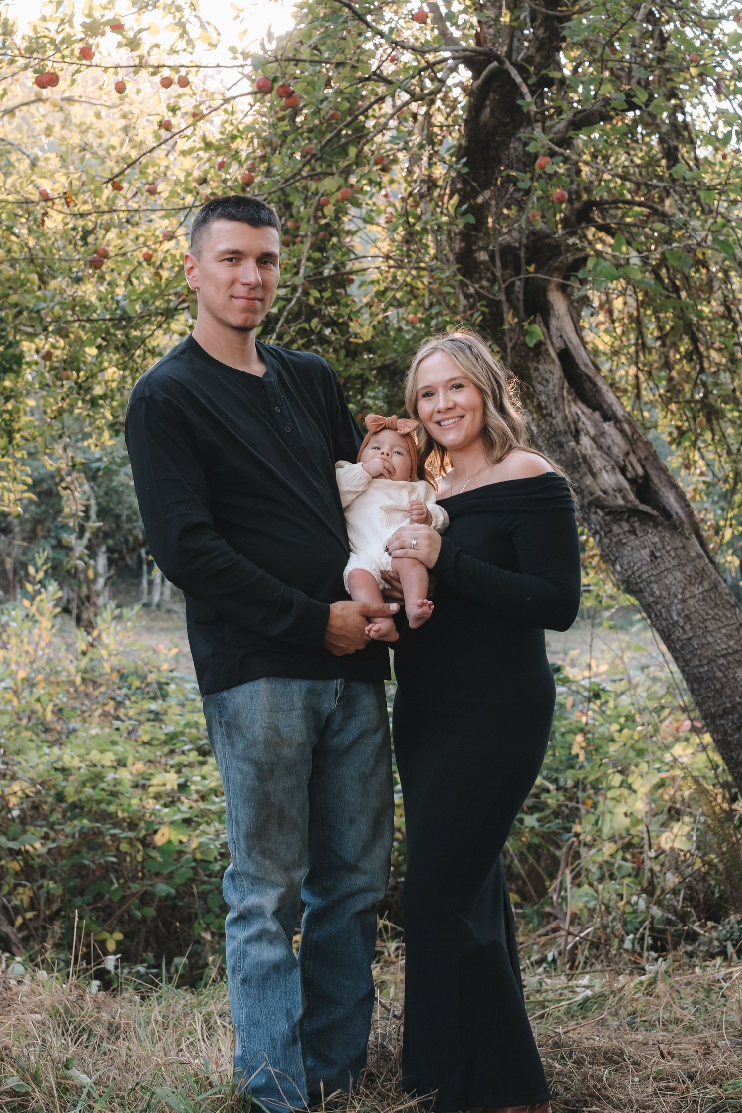 A couple embraces while holding a baby in an autumn outdoor setting with trees in the background.