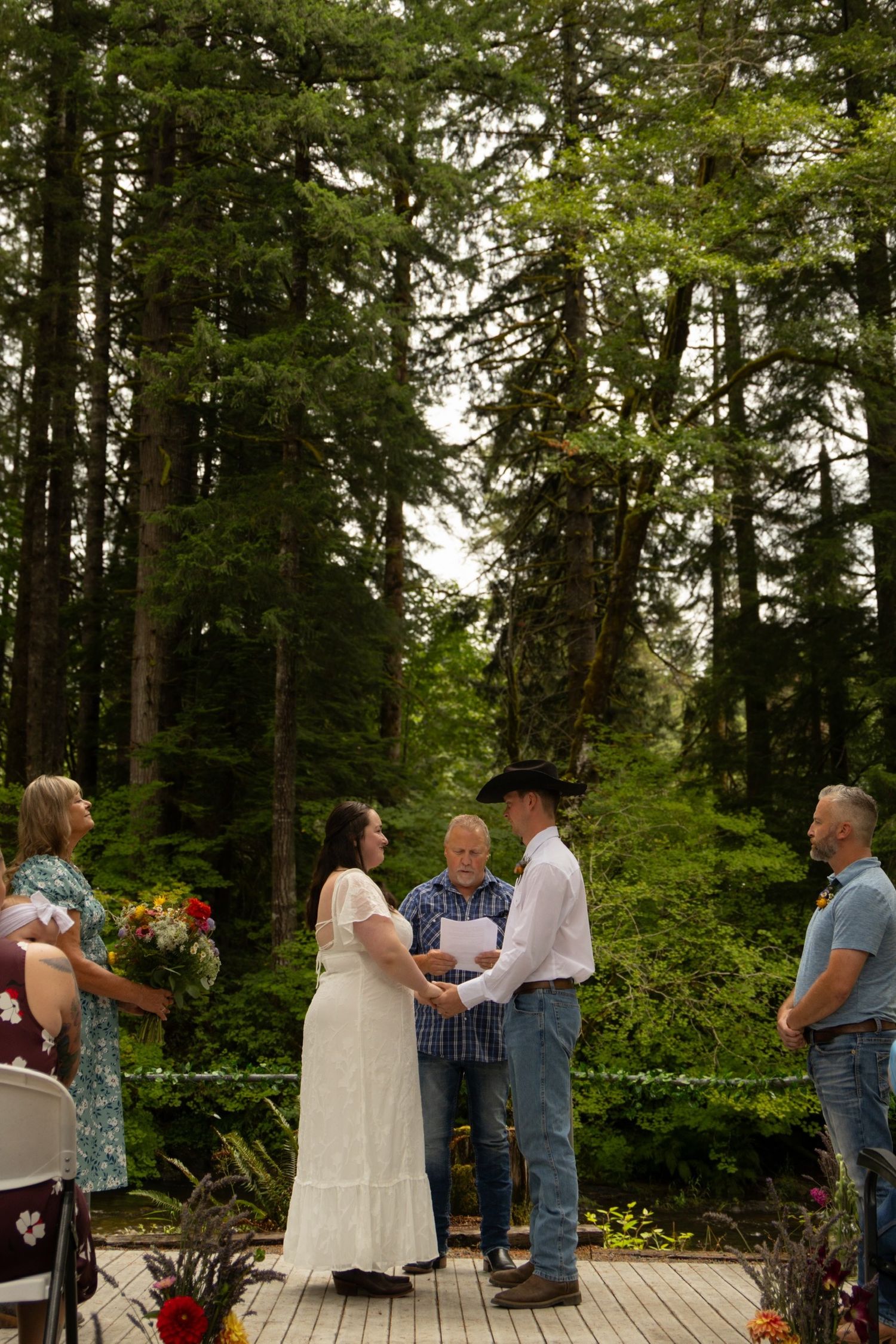 An intimate forest wedding ceremony taking place on a wooden deck with evergreen trees in the background.