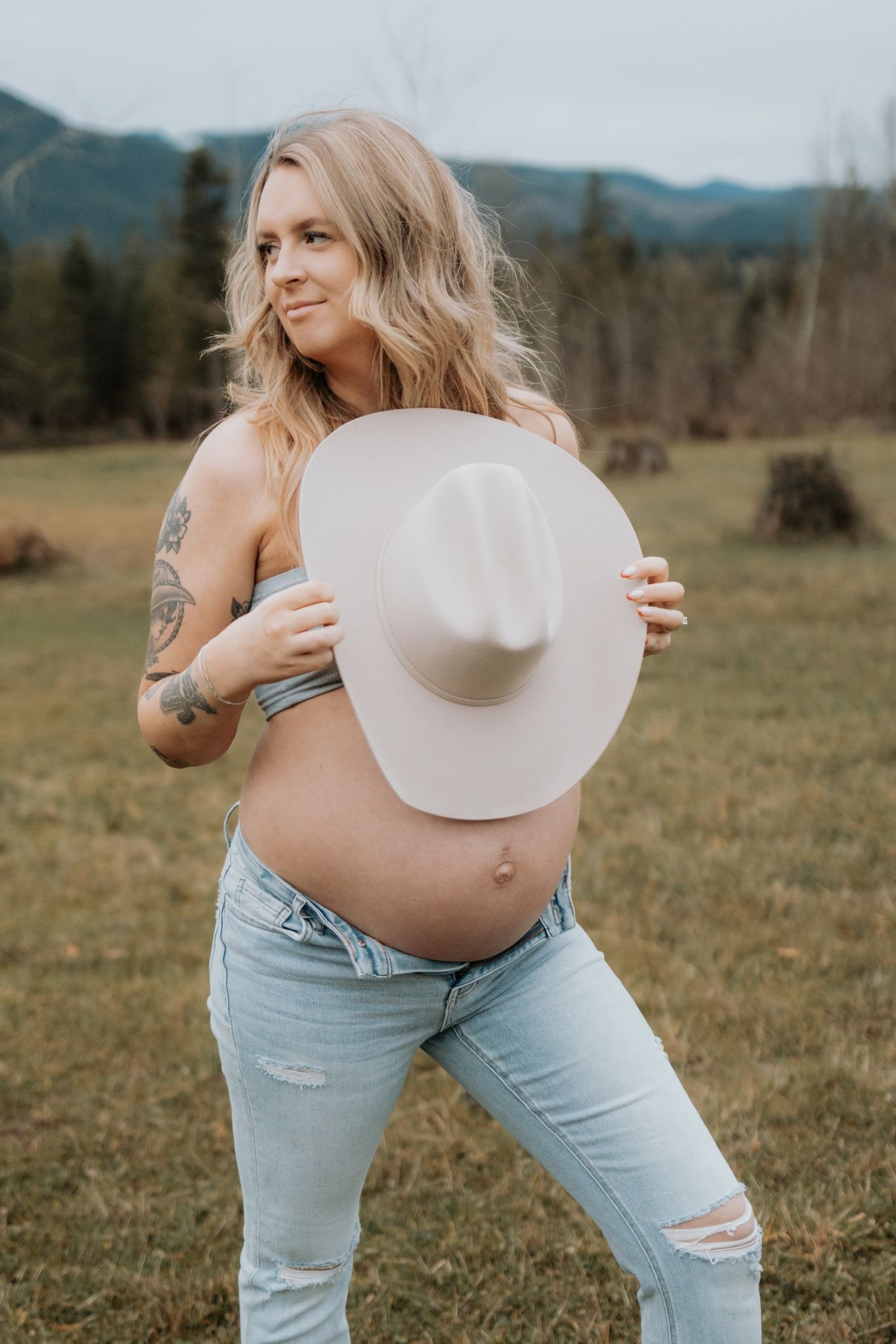 Country-style maternity portrait with white cowboy hat against mountain landscape.