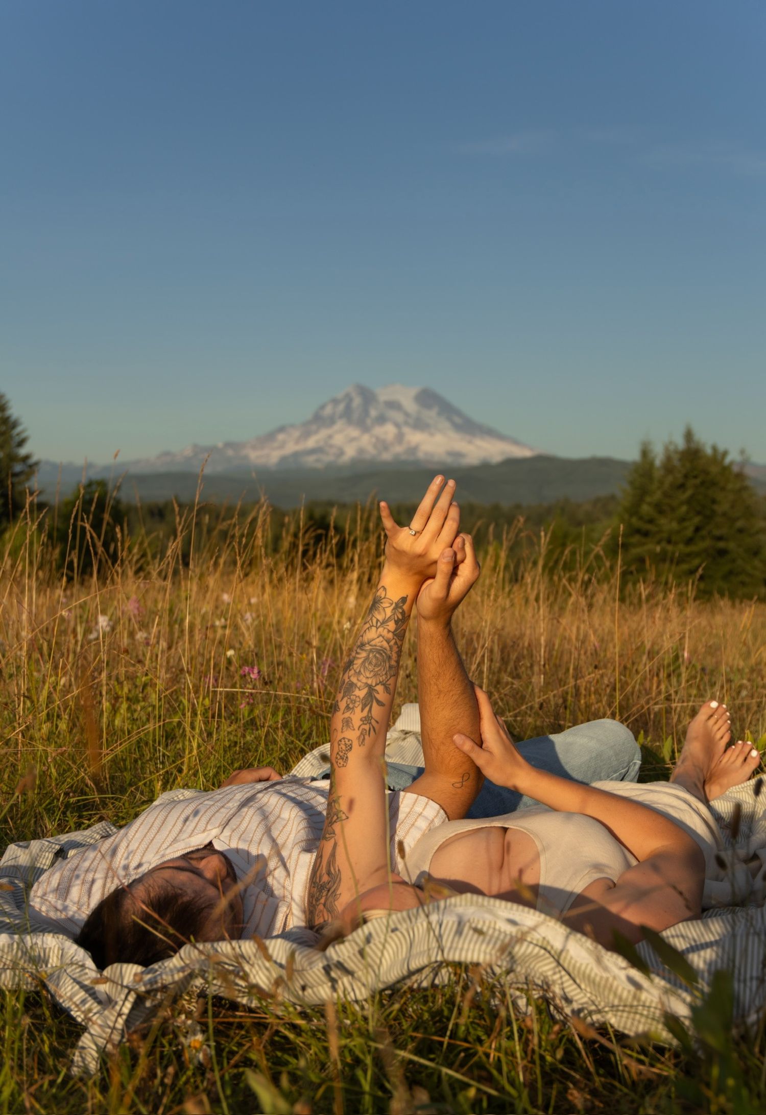 Two people lying on a blanket raising their hands together against a mountain backdrop.