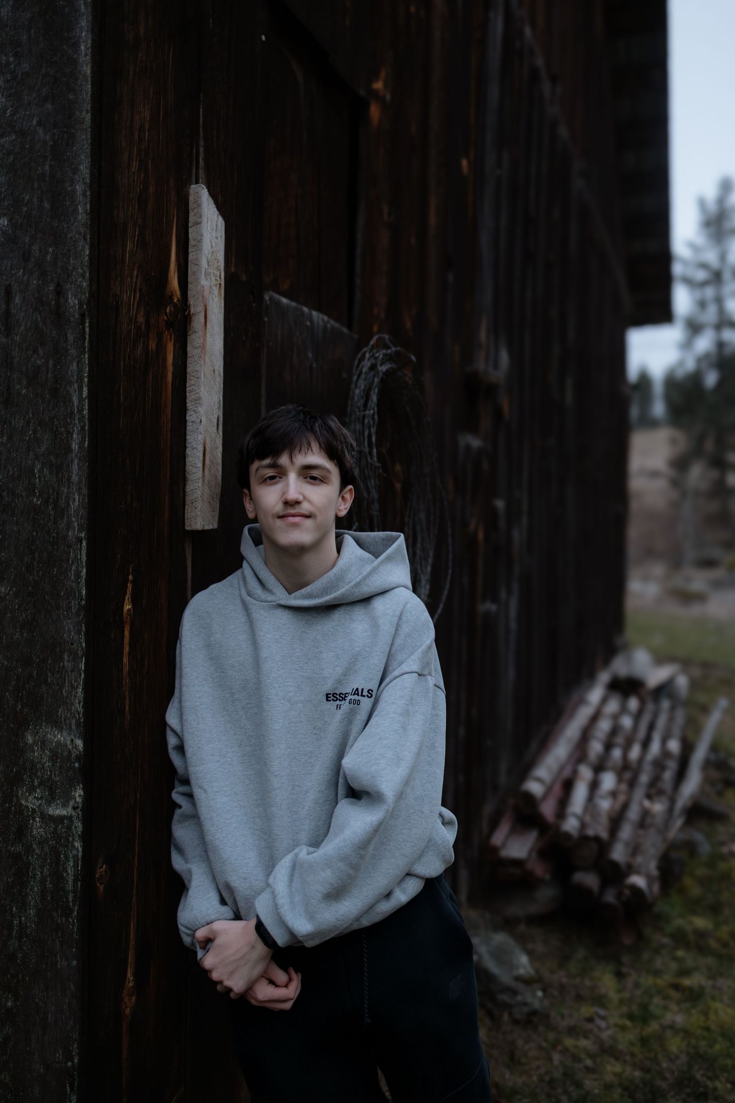 Person in a gray hoodie poses against dark wooden pillars in moody outdoor setting.