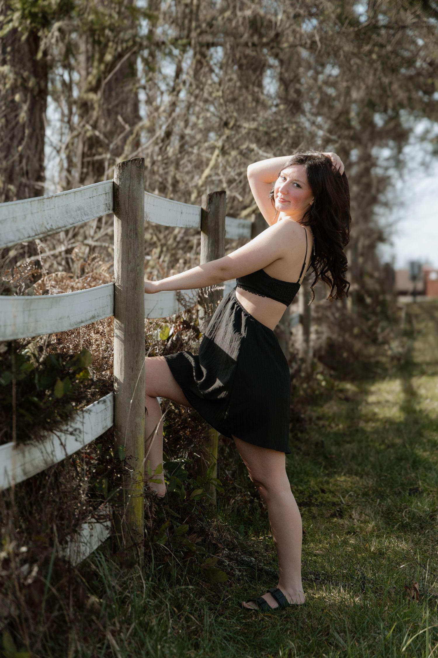 A dancer in a black dress performs graceful poses against a rustic wooden fence in a sunlit outdoor setting.