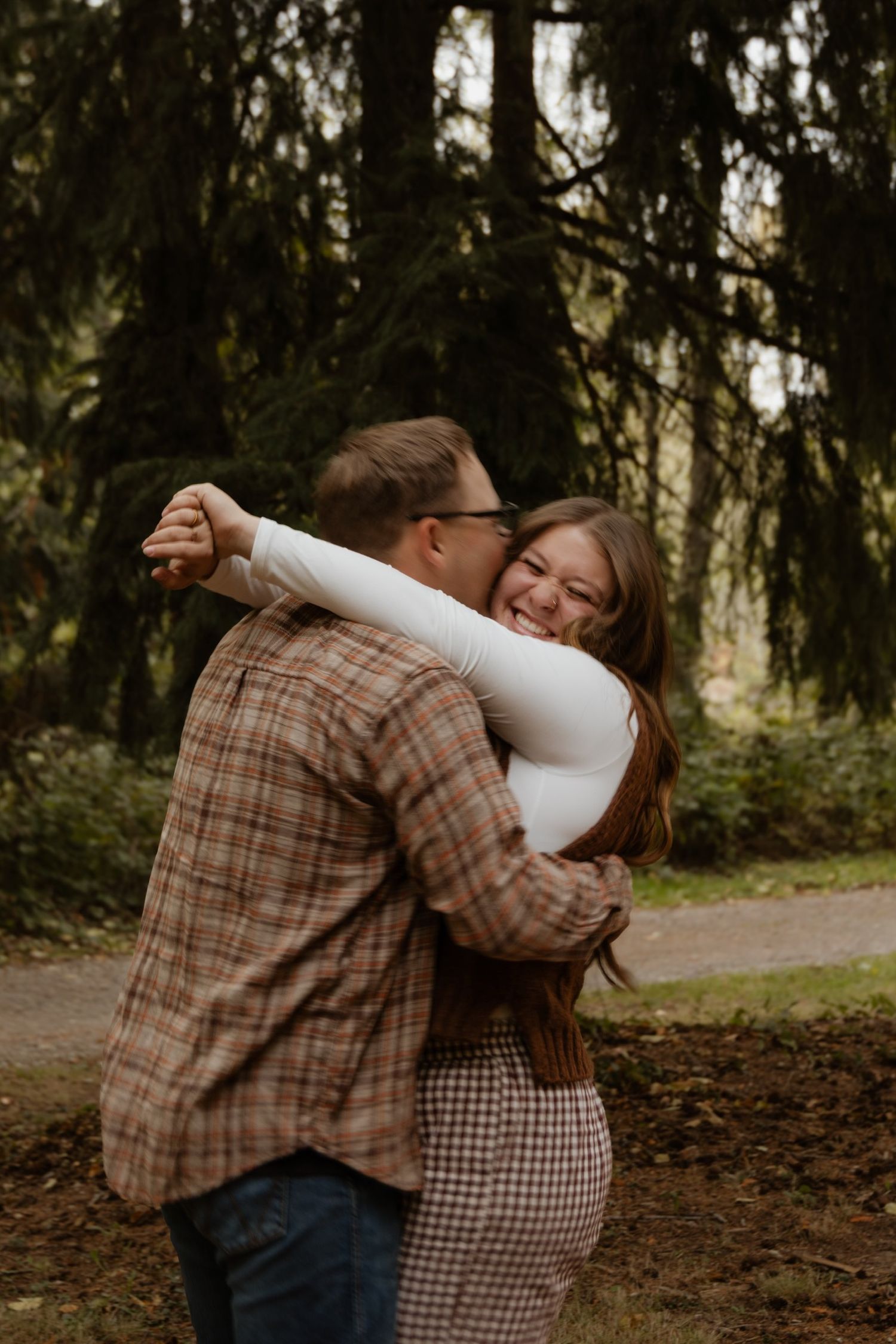 A couple embraces in a romantic moment among tall evergreen trees in a forest setting.