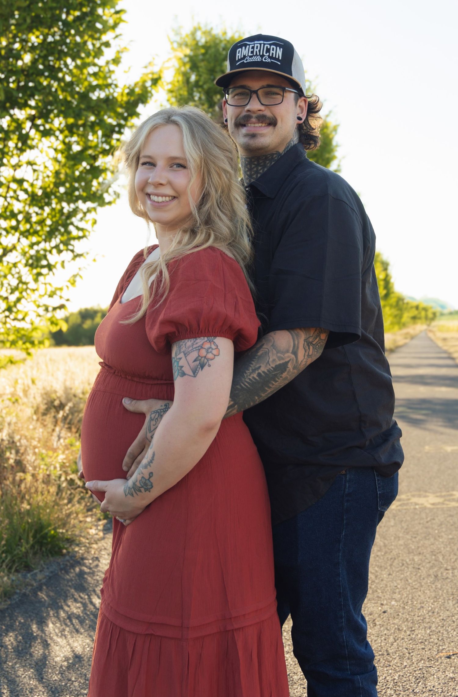 A couple poses outdoors in sunlight, with a red dress and casual attire against a nature backdrop.