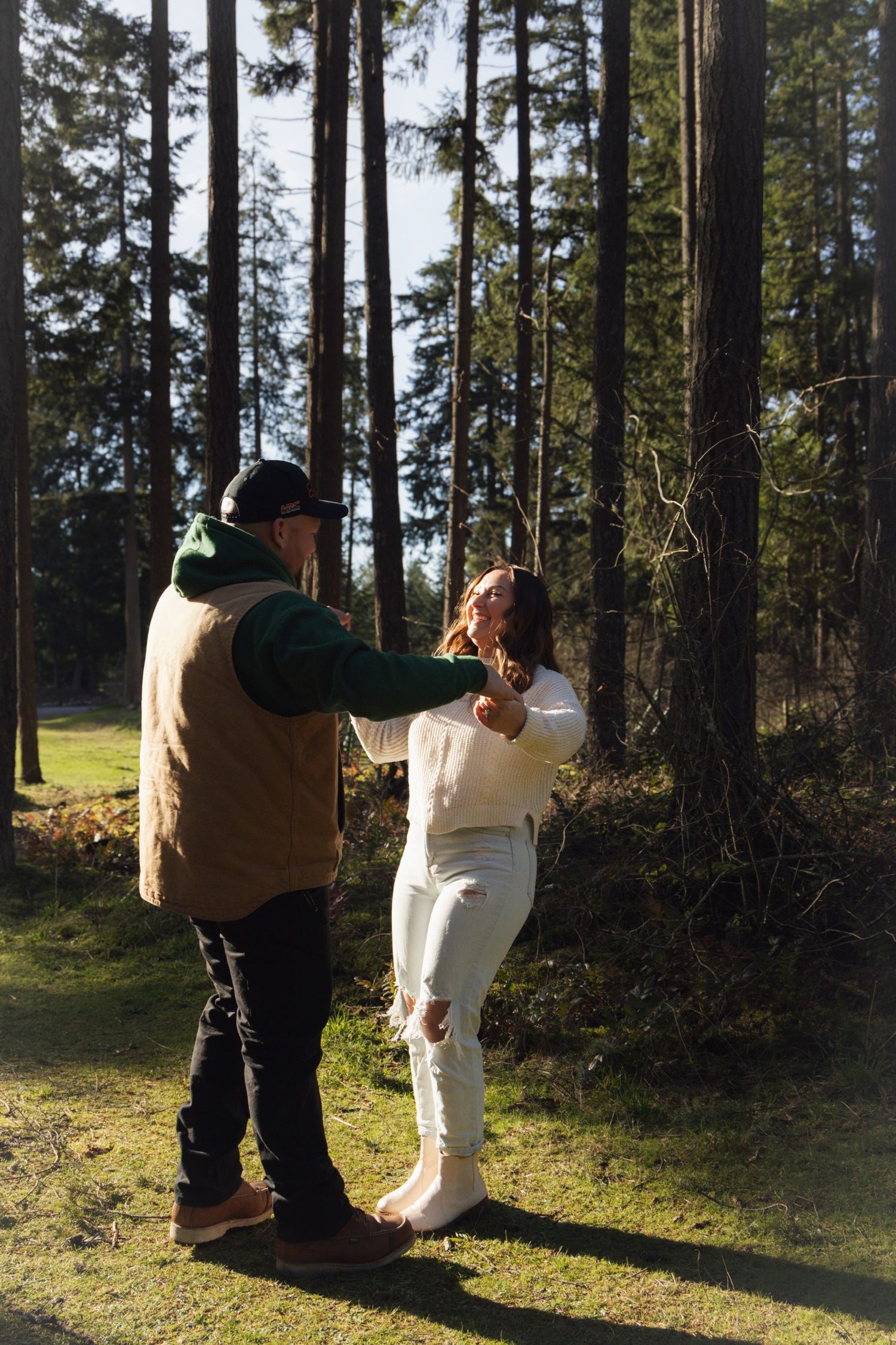 A couple walking hand in hand through a sunlit forest with tall evergreen trees in the background.