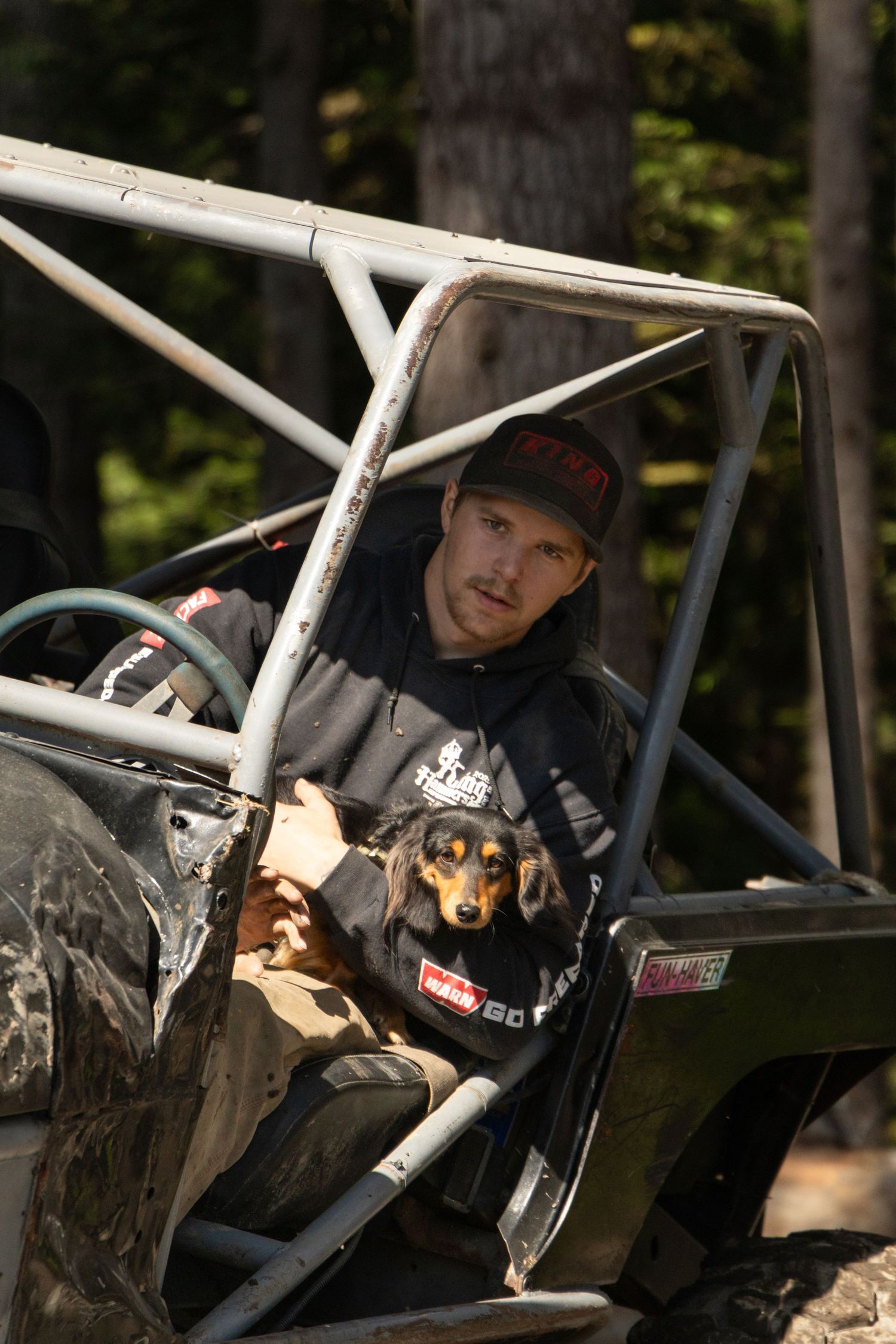 A person sits in a dark colored off-road vehicle with a roll cage in a wooded outdoor setting.