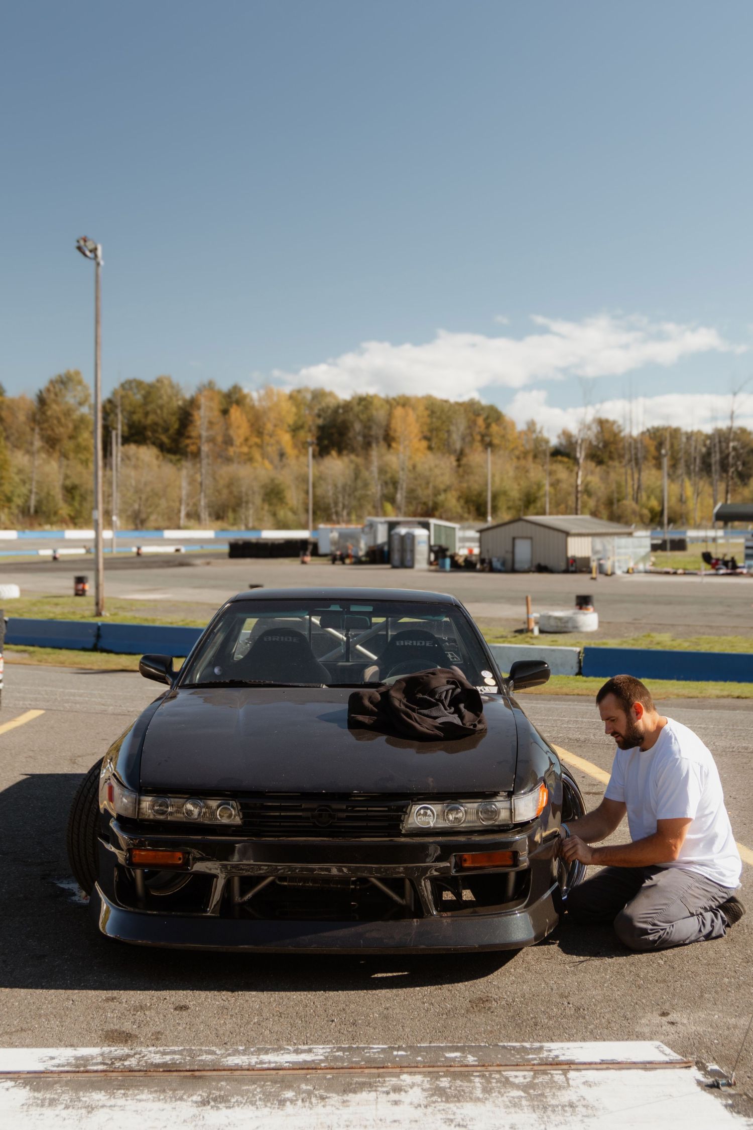 Black Nissan Silvia S13 drift car parked in a lot with autumn trees and trailers in the background.