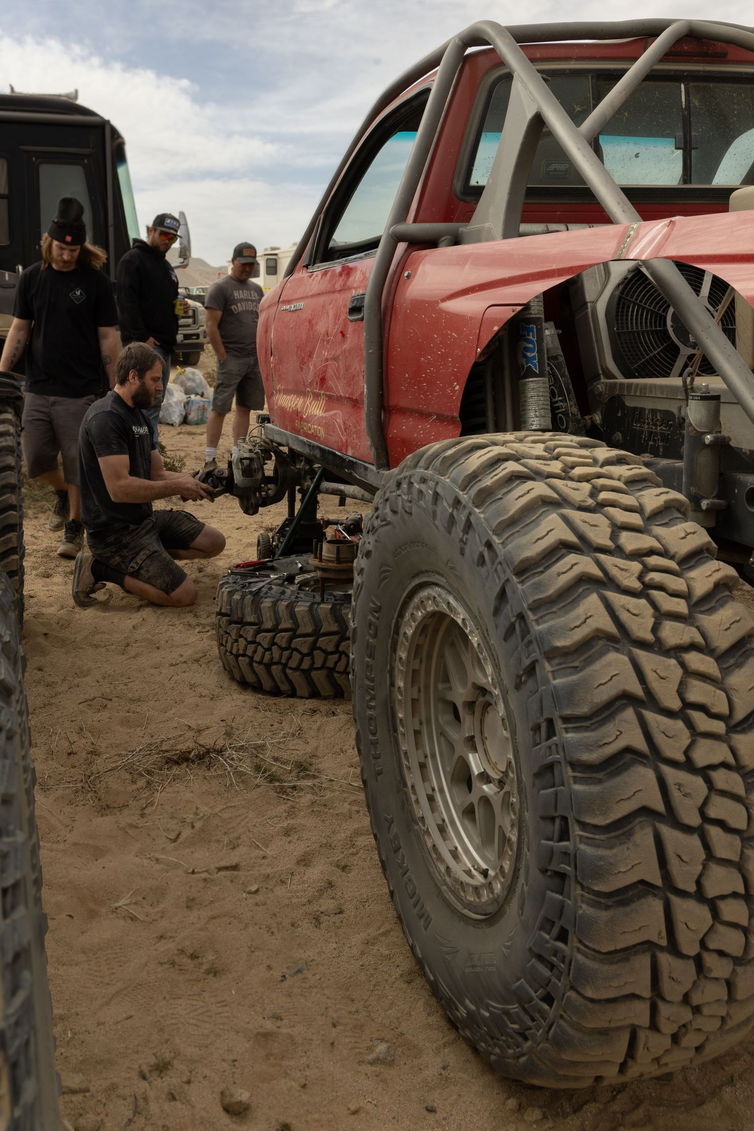 A red off-road truck with large rugged tires and roll cage being worked on in sandy terrain.