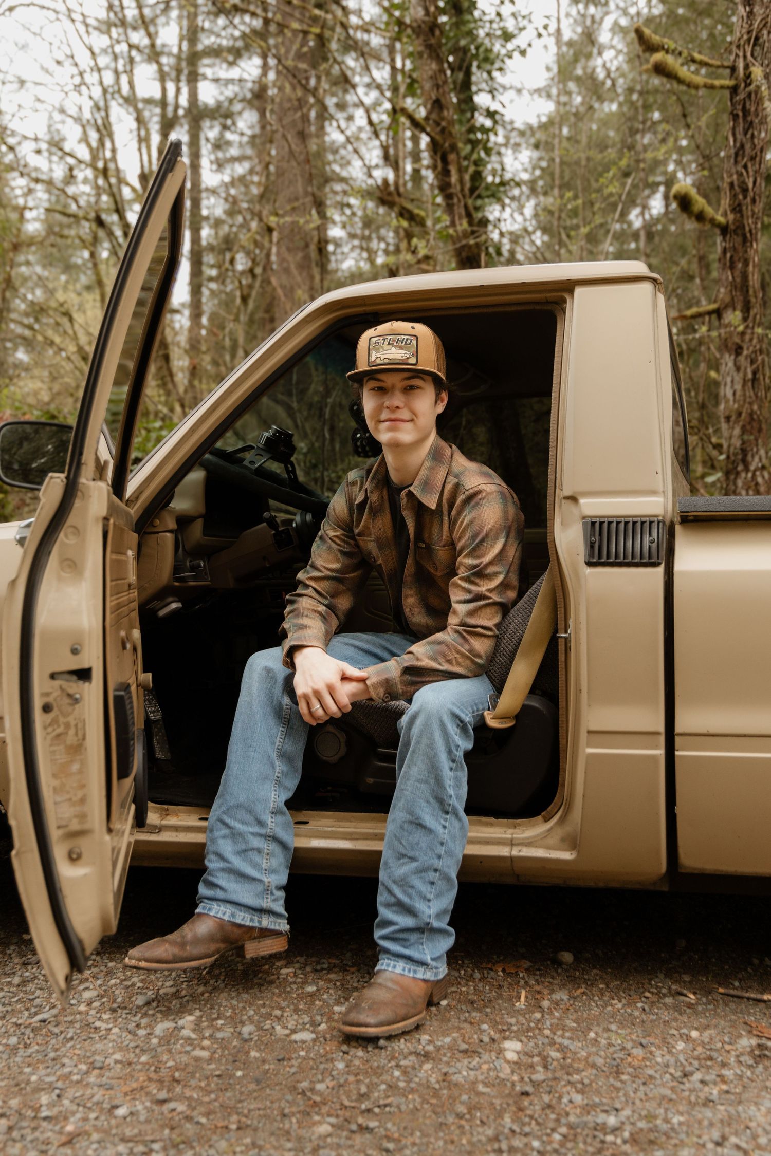 Young man in flannel shirt and trucker hat sitting in open door of tan pickup truck in forest setting.