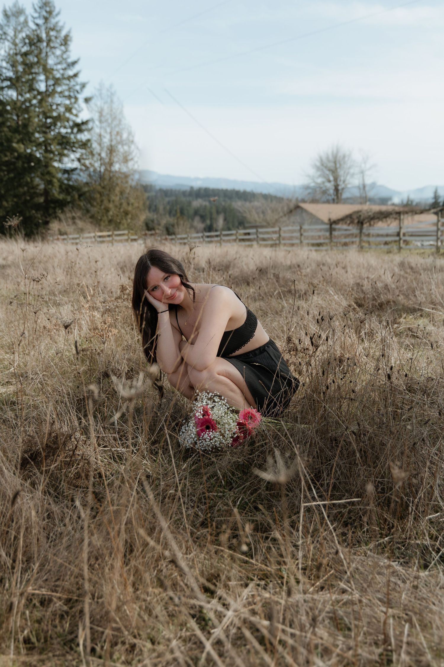 A figure in black attire crouches in a field of tall grass with mountains and a wooden fence in the background.
