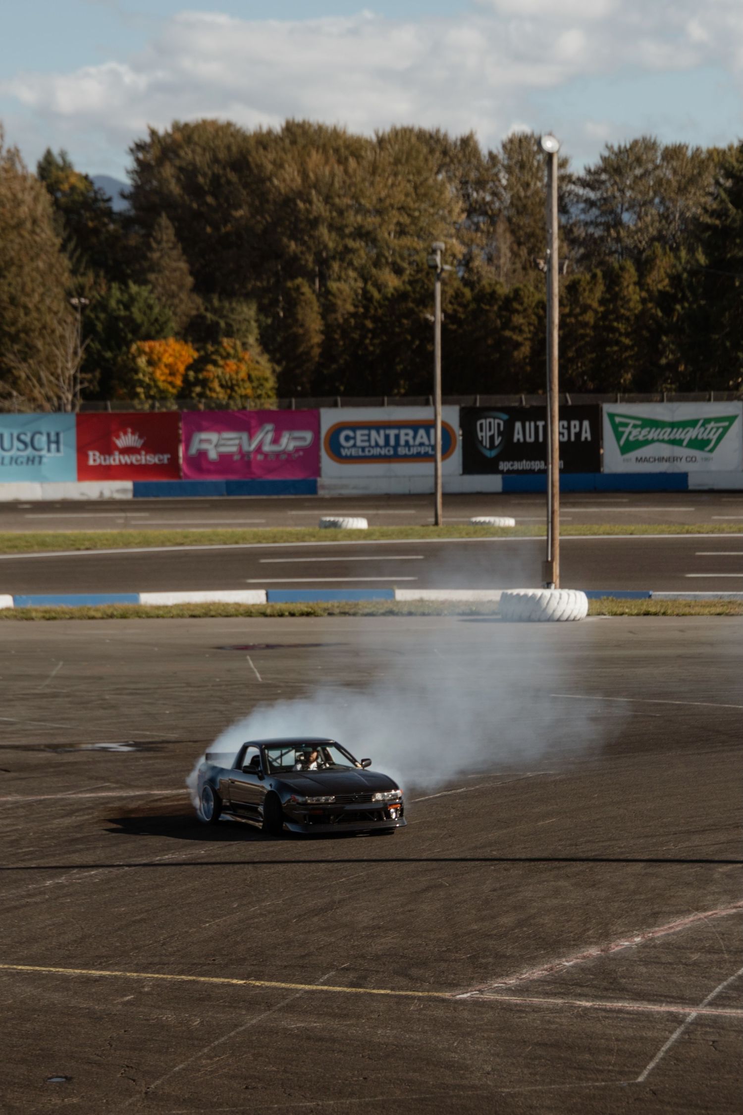 A black sports car drifts and creates smoke while performing donuts in an empty parking lot with sponsor banners visible.