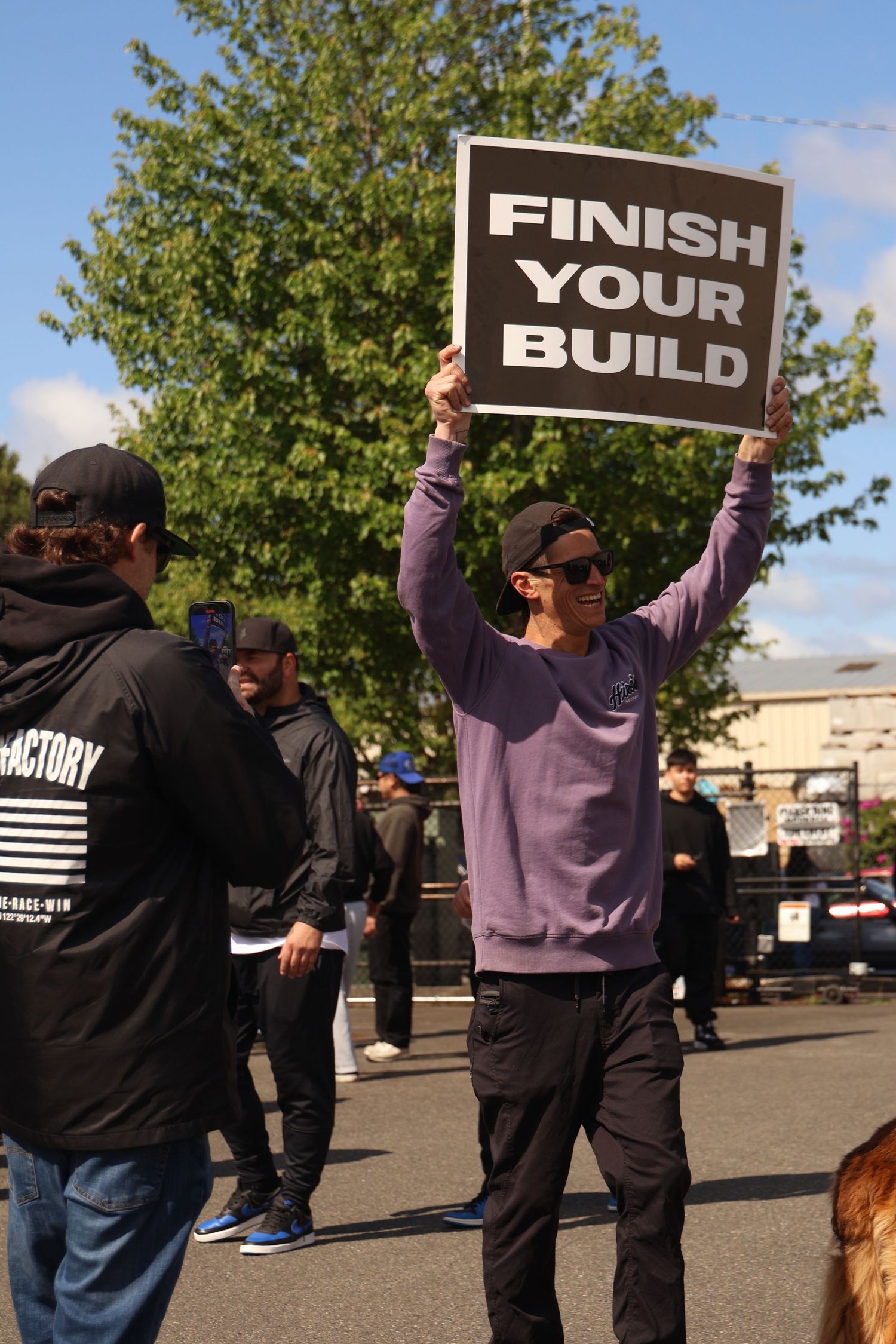 Two people hold up black signs with white text reading 'FINISH YOUR BUILD' while standing in a parking lot.